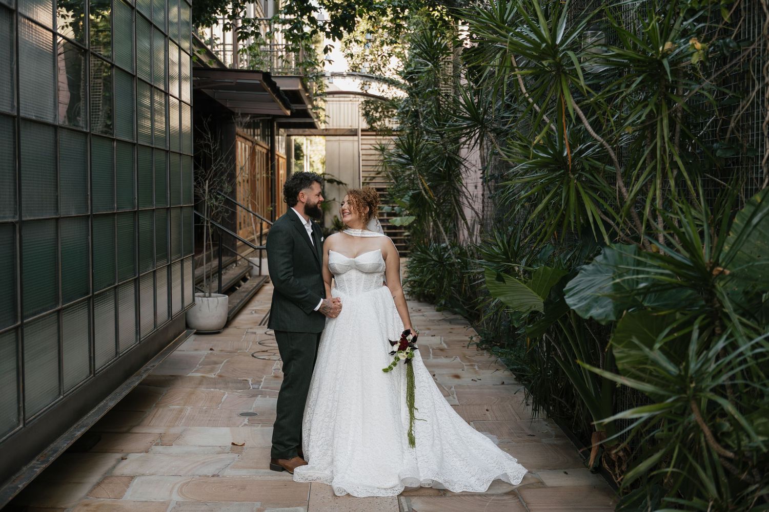 Bride and groom share a romantic moment in a lush garden walkway, bride holding a bouquet in a white gown.