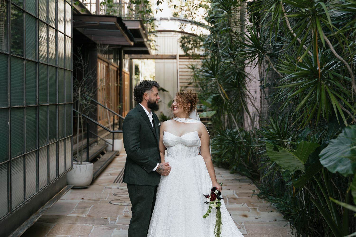 Bride and groom holding hands in a lush courtyard, bride in white ballgown, groom in dark suit.