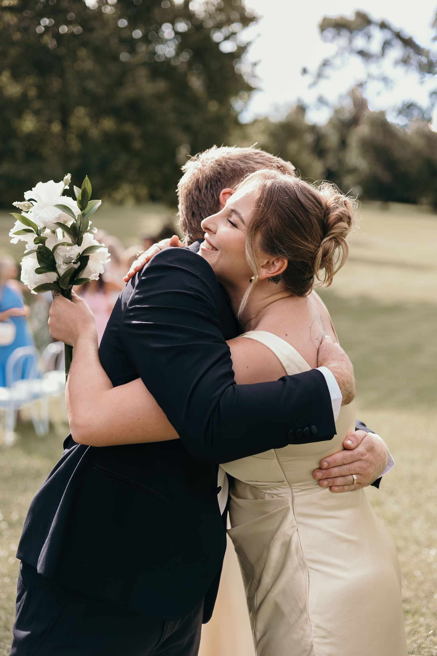 Bride and groom share a warm embrace outdoors during wedding ceremony, bride holding white floral bouquet.