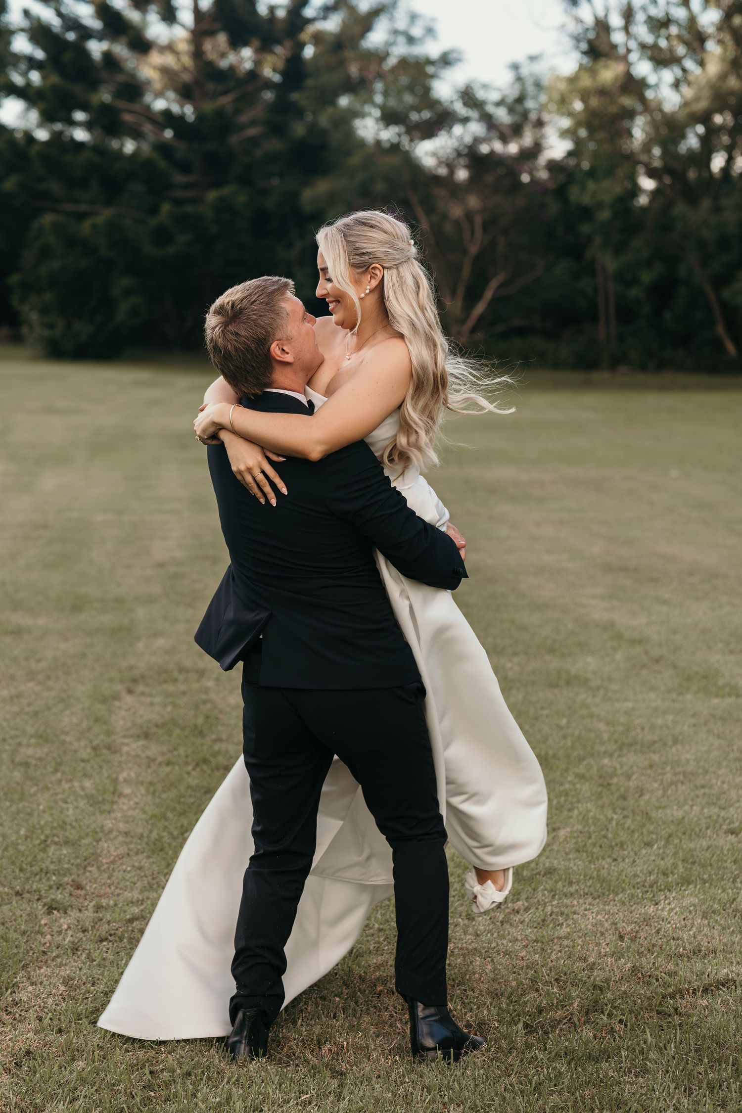 Groom lifting bride in white gown on green lawn, both laughing joyfully during outdoor wedding photo session.