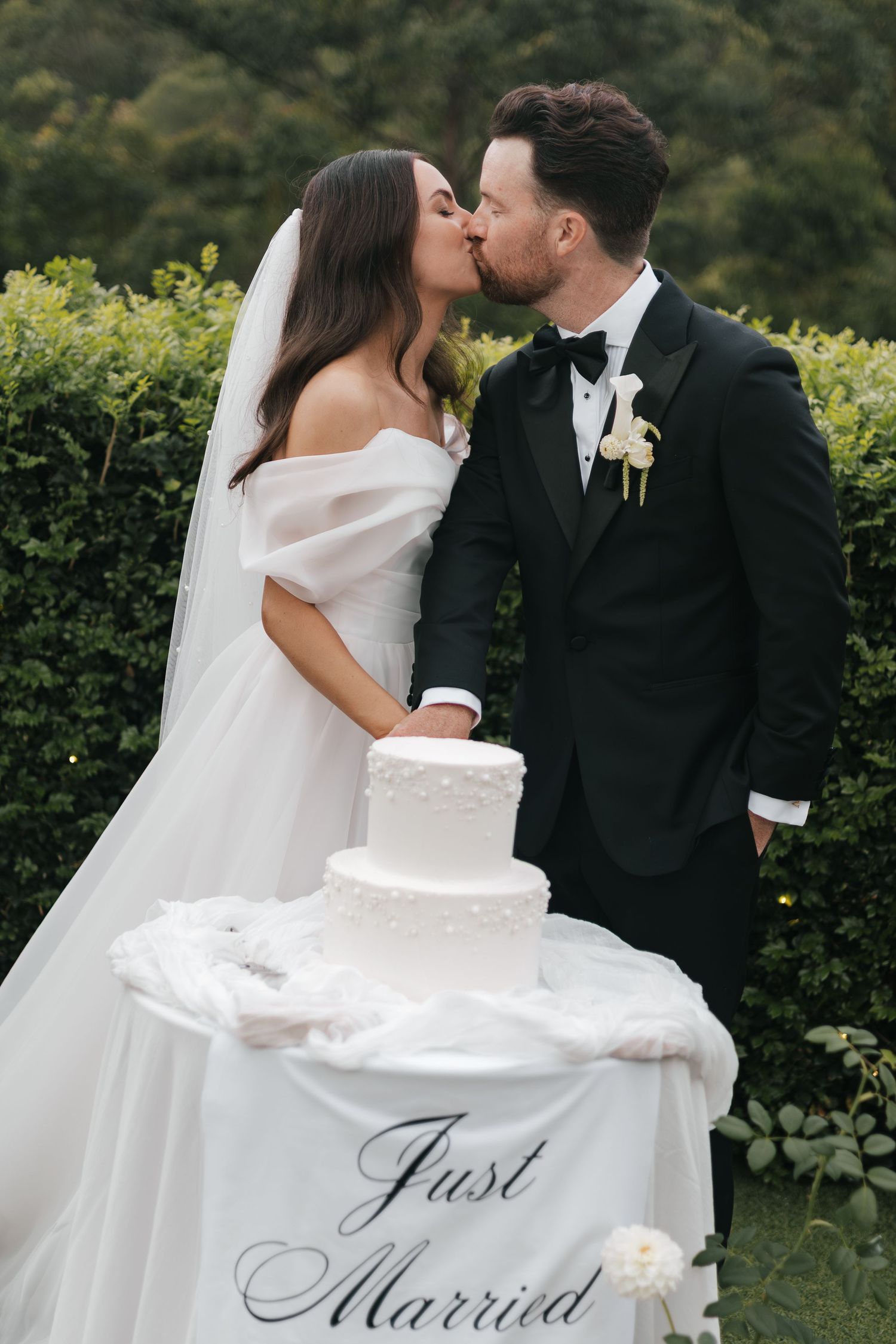 Bride and groom kissing behind a white wedding cake on a Just Married table outdoors.