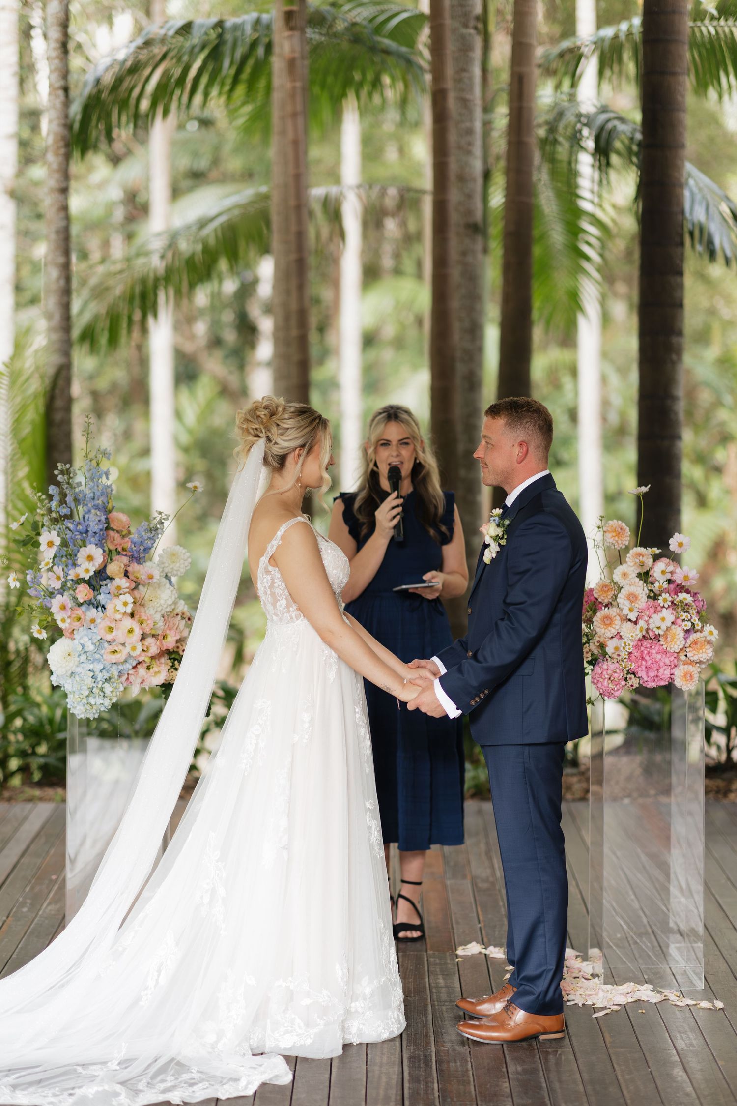 Bride and groom exchange vows outdoors on wooden deck surrounded by lush tropical trees and colorful floral arrangements.