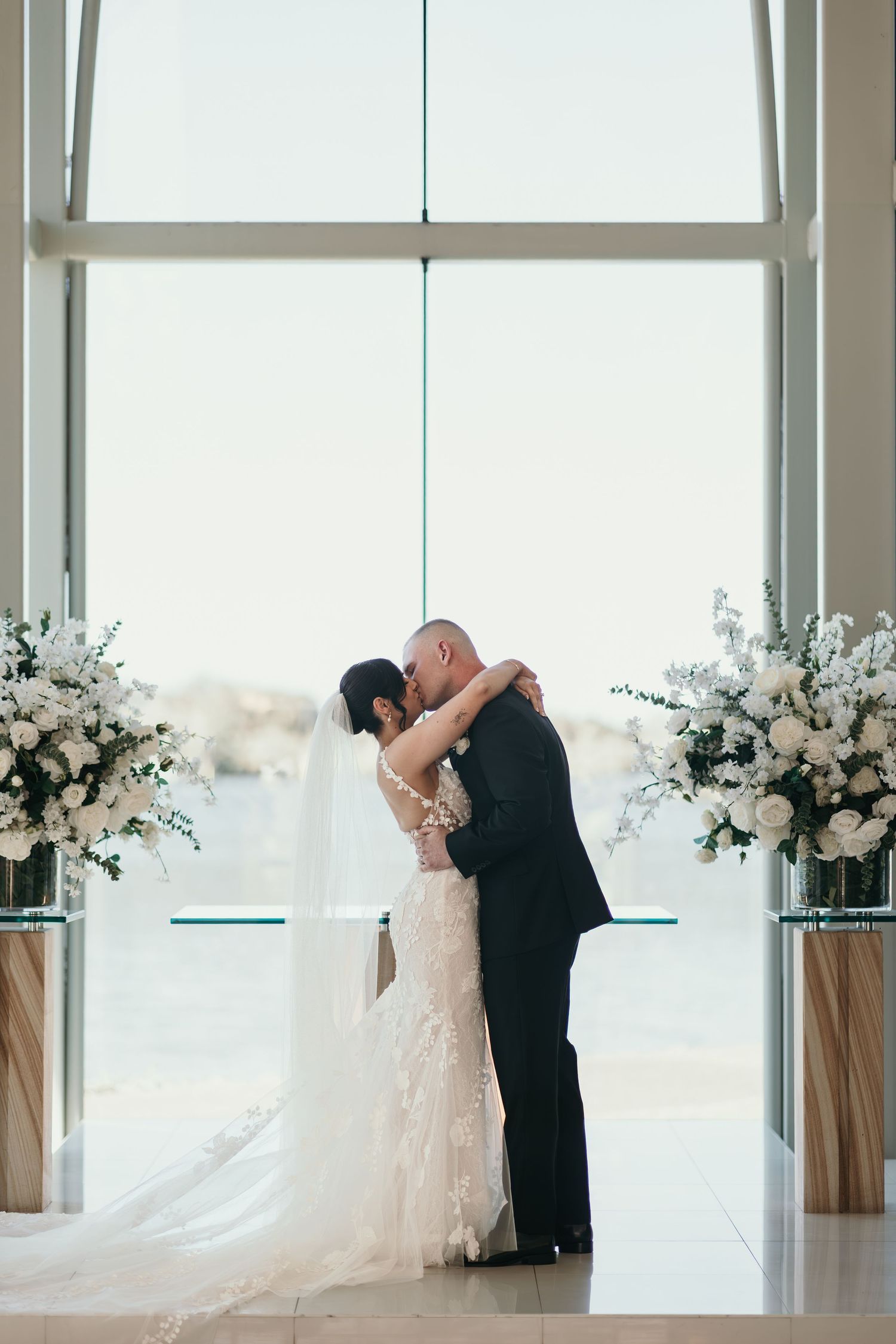 A couple shares an intimate embrace during their wedding ceremony in a bright modern venue with white floral arrangements.