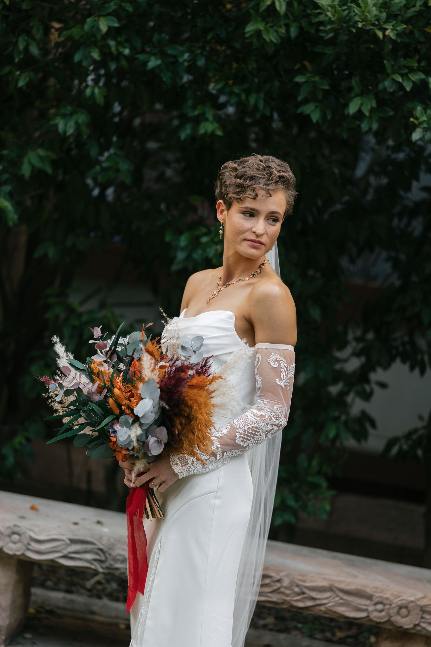 Bride in strapless white gown with lace sleeves holds colorful bouquet outdoors near green foliage.