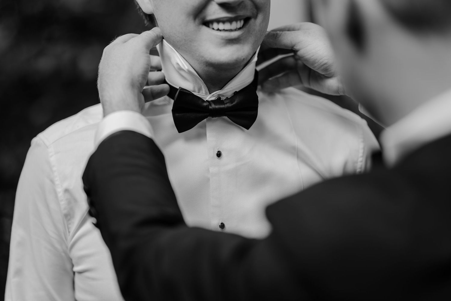 Black and white photo of a groom smiling as someone adjusts his bow tie before the wedding ceremony.