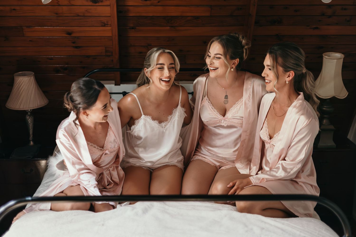 Four bridesmaids in pink satin robes laughing together on a bed in a rustic wooden cabin bedroom.