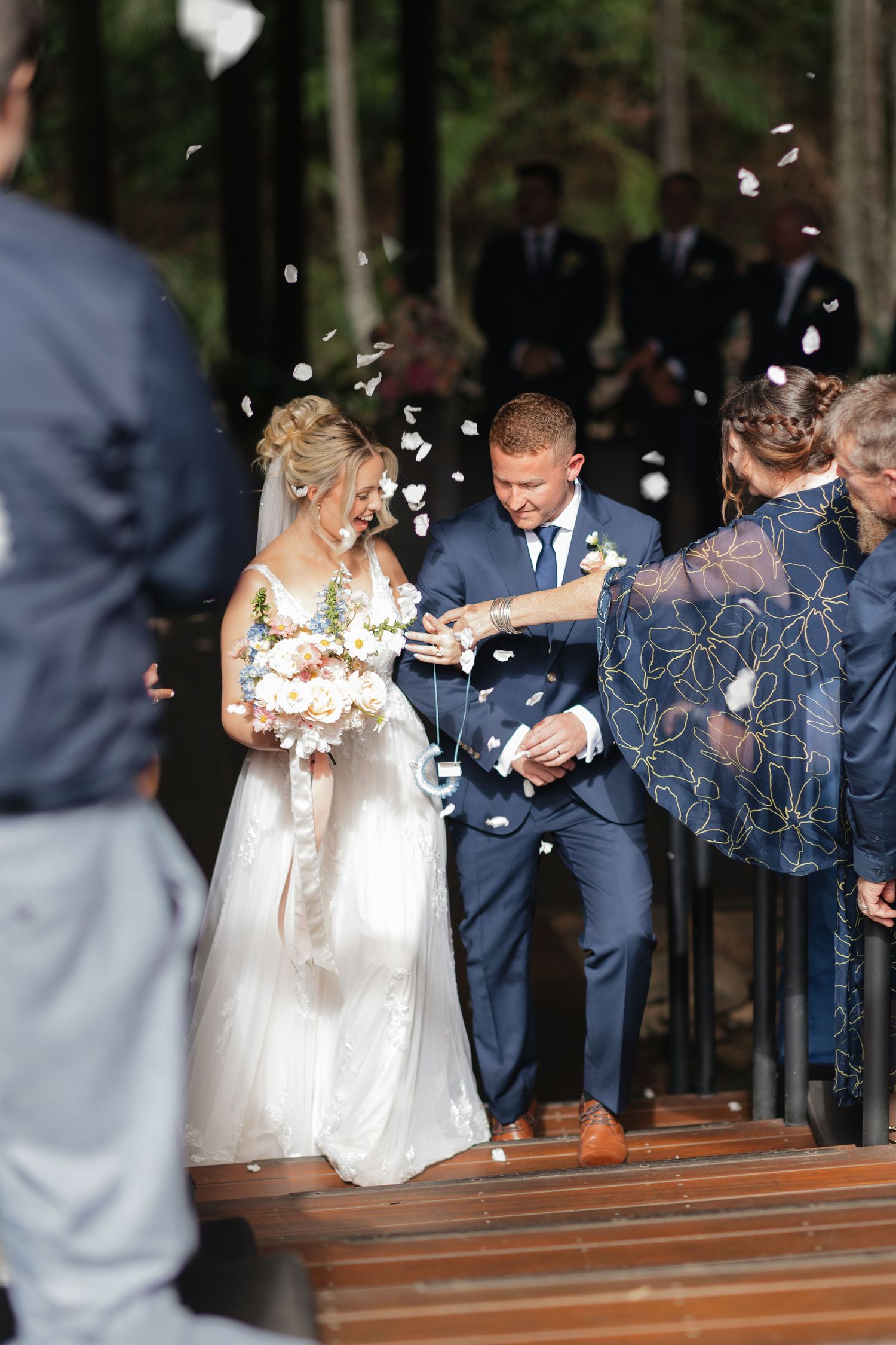 Bride and groom walk down aisle as guests toss flower petals at outdoor forest wedding ceremony.