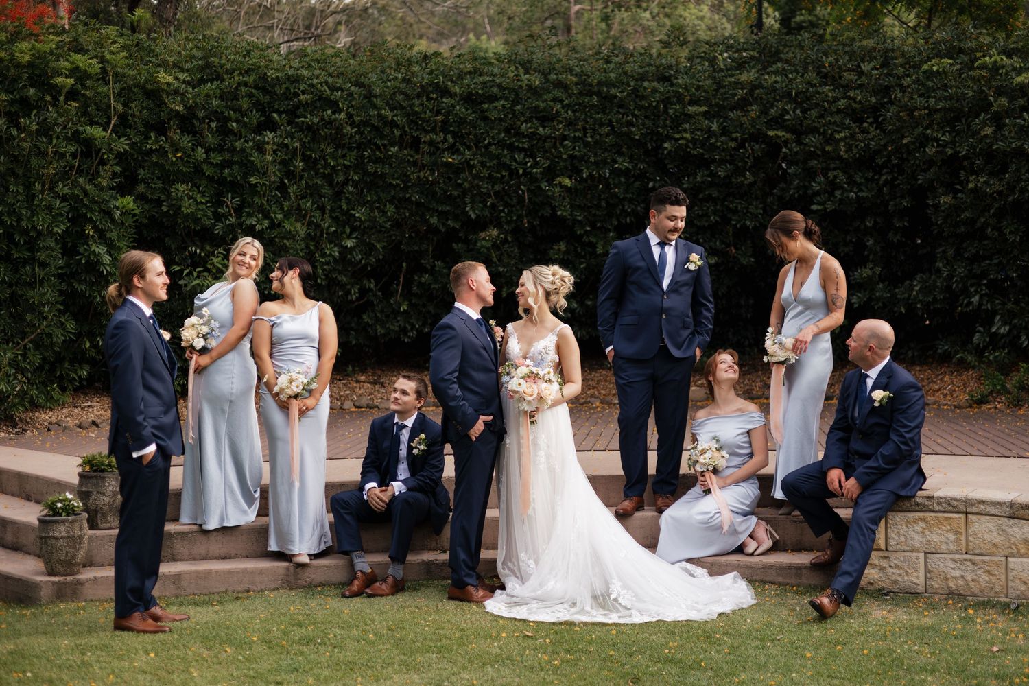 Wedding party poses outdoors, bride and groom centered, bridesmaids in silver gowns, groomsmen in navy suits, lush greenery backdrop.