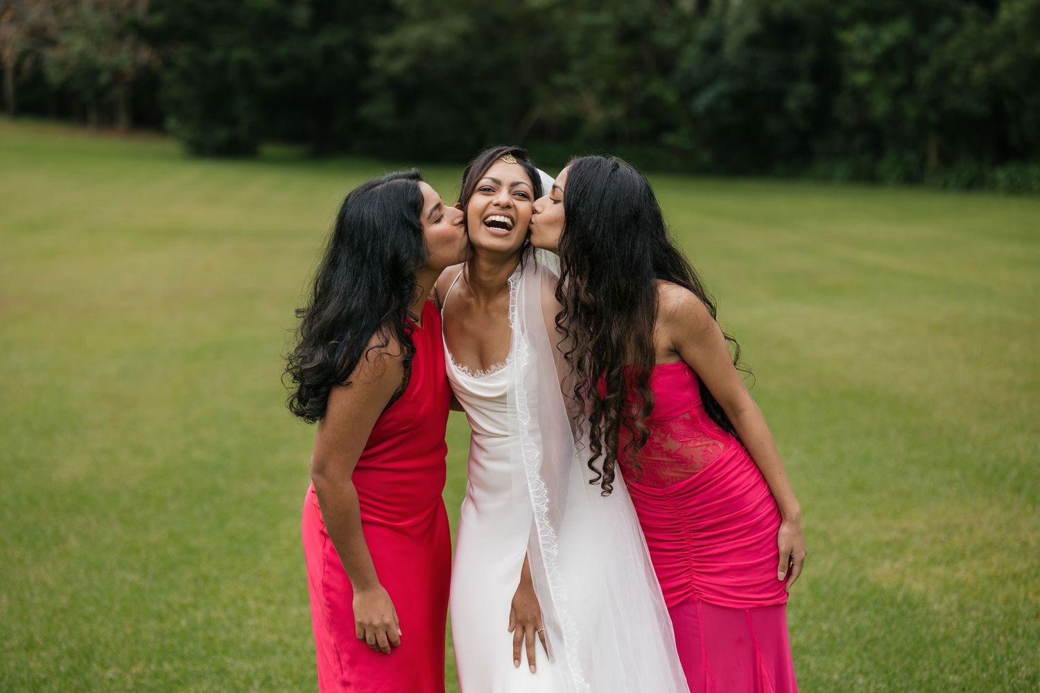 Bride in white gown laughing as two bridesmaids in pink dresses kiss her cheeks outdoors on green lawn.