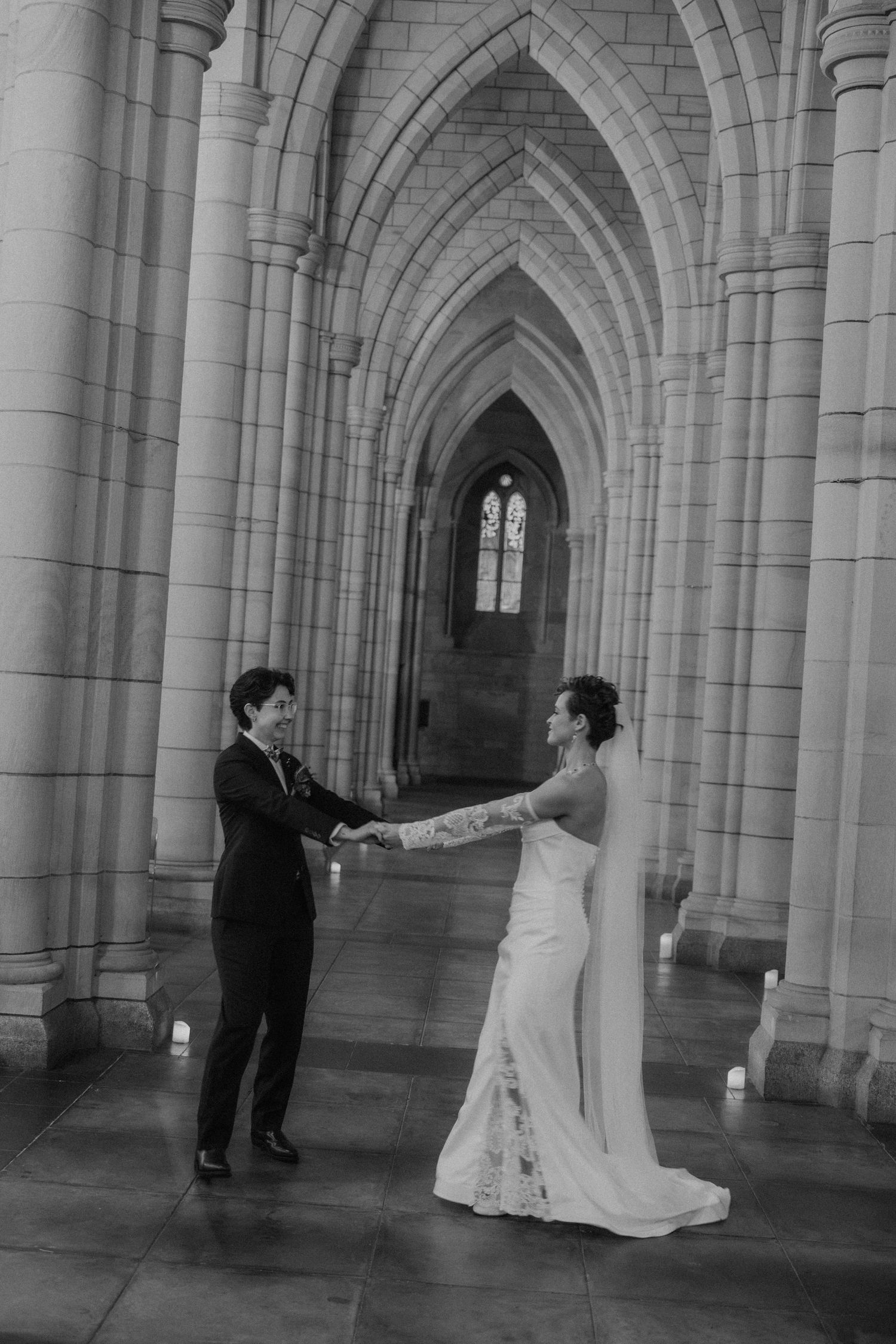 Black and white photo of a bride and groom holding hands in a Gothic cathedral archway with candlelit stone columns.