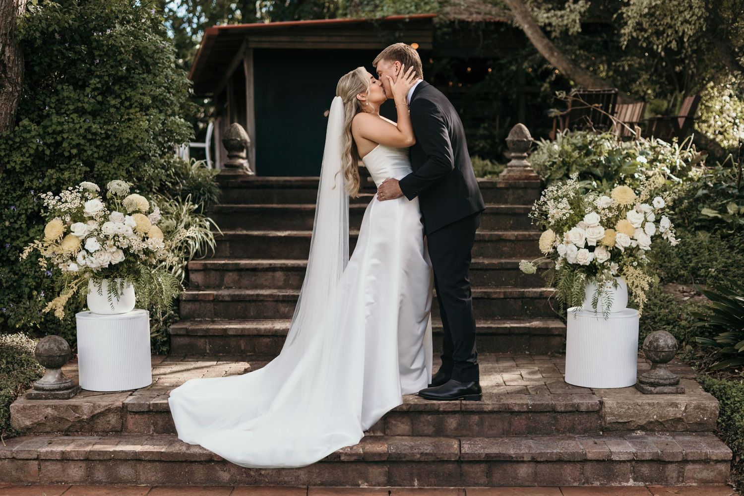 Bride and groom sharing a kiss on stone steps surrounded by white floral arrangements at an outdoor wedding ceremony.