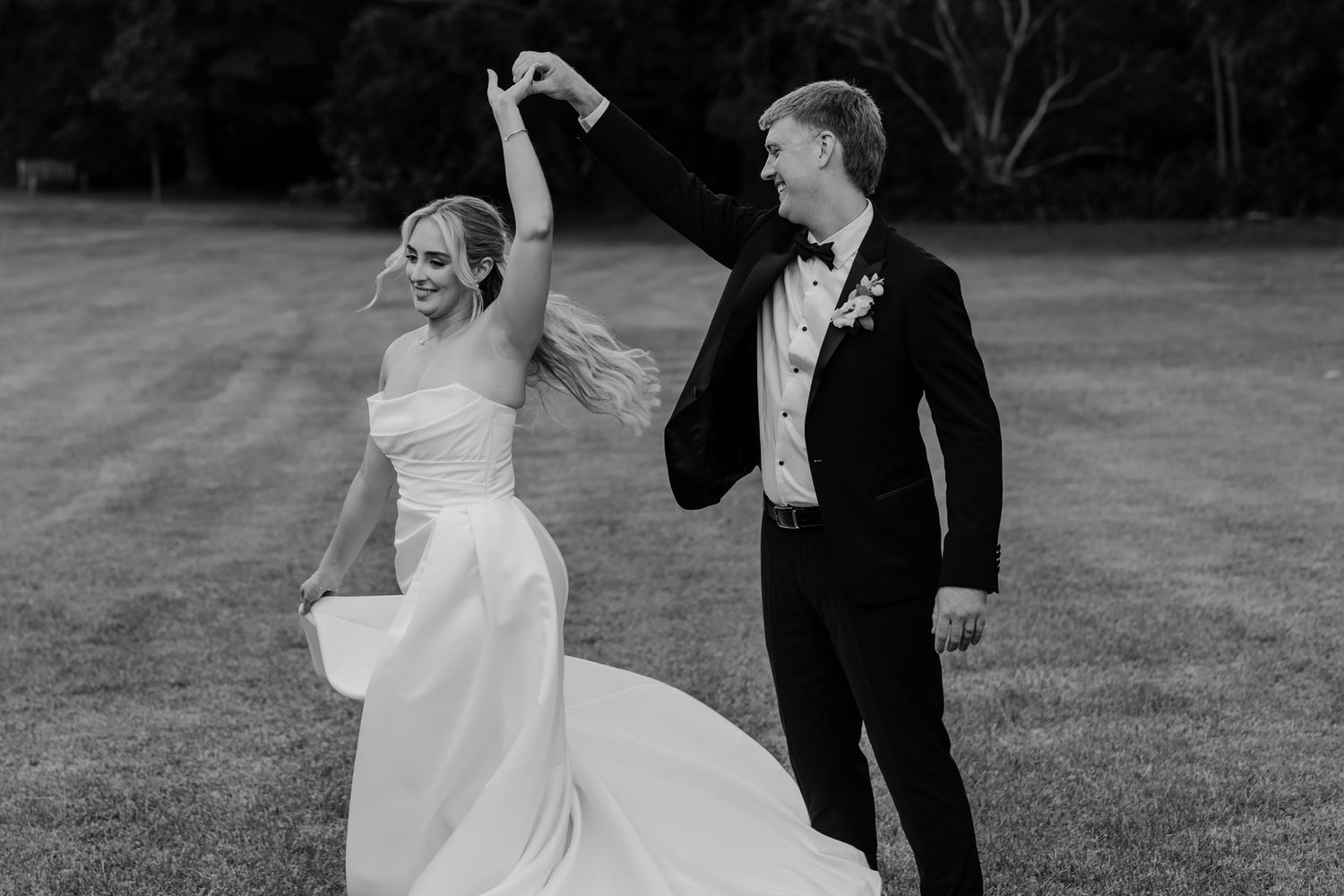 Black and white photo of a bride and groom dancing outdoors on a lawn, groom twirling bride in wedding attire.