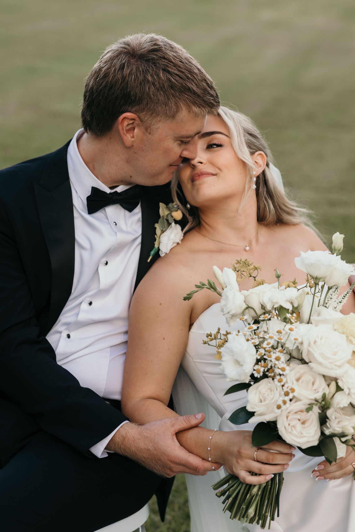 Groom in black tuxedo kisses bride holding white rose bouquet outdoors on their wedding day.