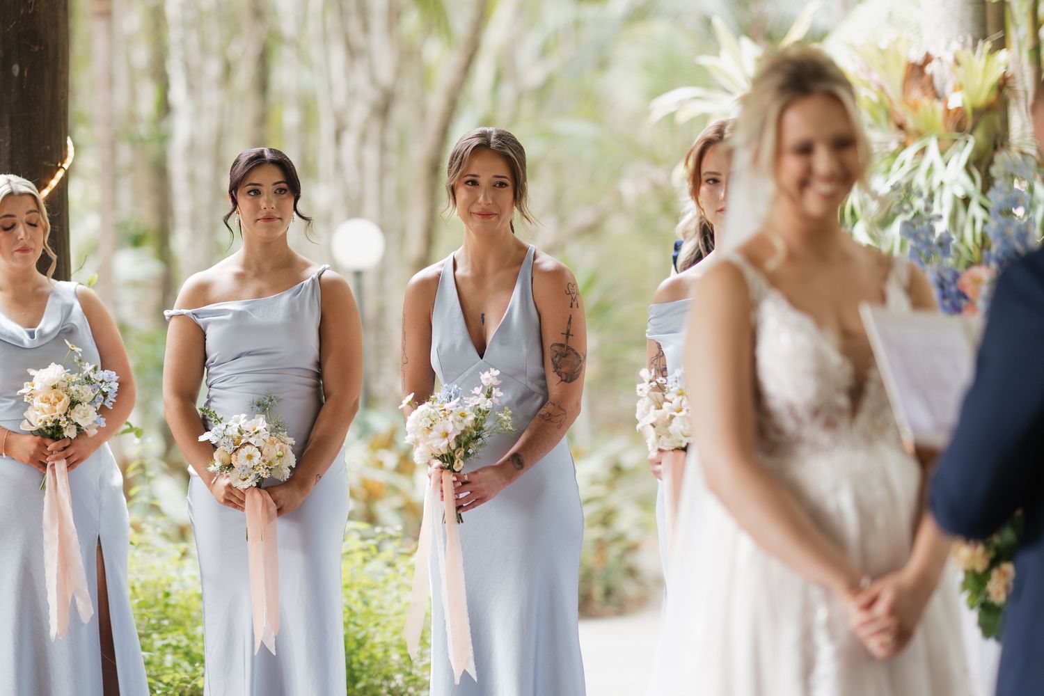 Bridesmaids in light blue dresses holding bouquets stand attentively as bride in lace gown faces the altar at outdoor wedding.