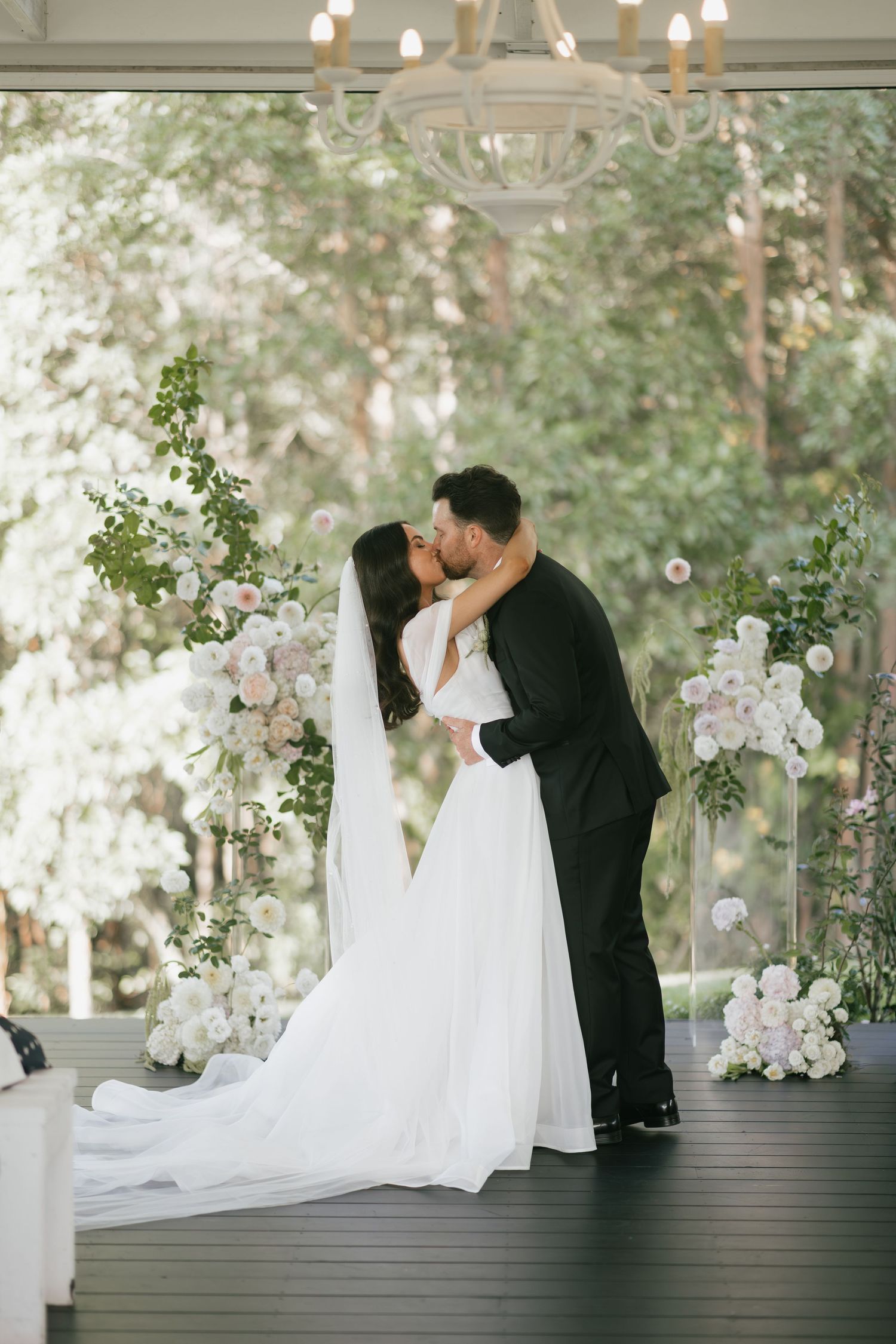 Bride and groom share first kiss at elegant wedding ceremony with white floral arrangements and greenery backdrop.