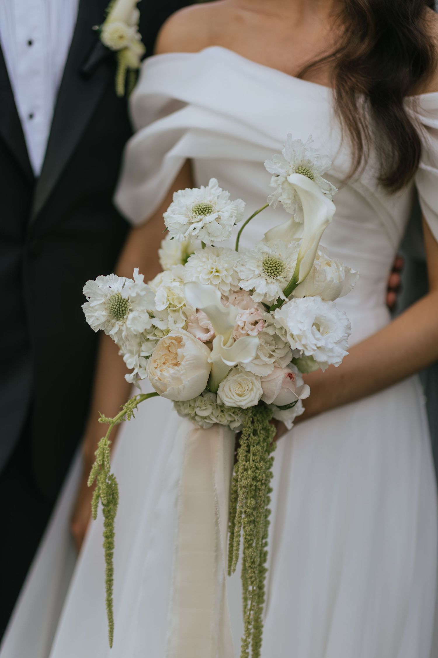 Bride holding a lush white and blush wedding bouquet with cascading greenery, standing beside groom in black tuxedo.