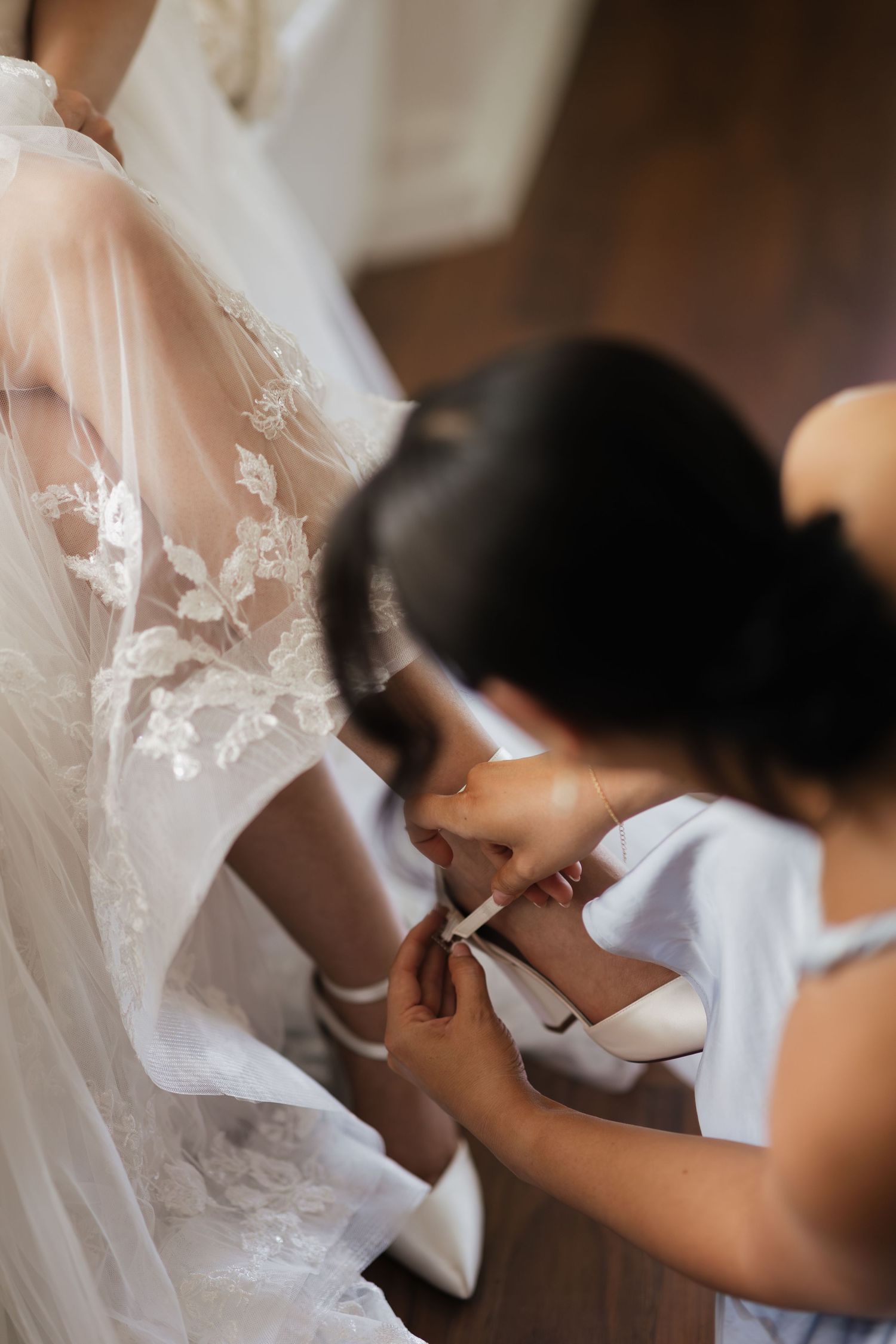 Bridesmaid helping bride put on white heeled shoes, with lace wedding dress detail in soft natural light.