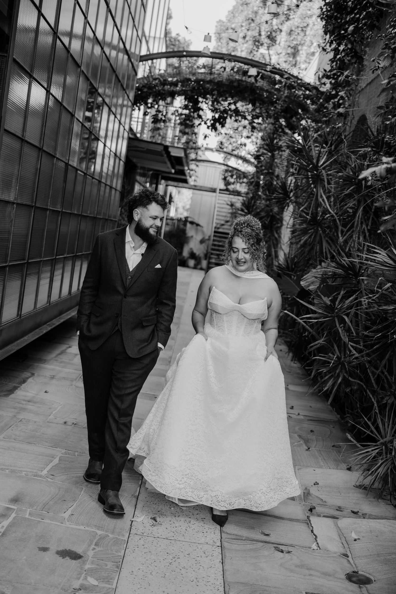 Black and white photo of a bride and groom walking together through a lush garden pathway on their wedding day.