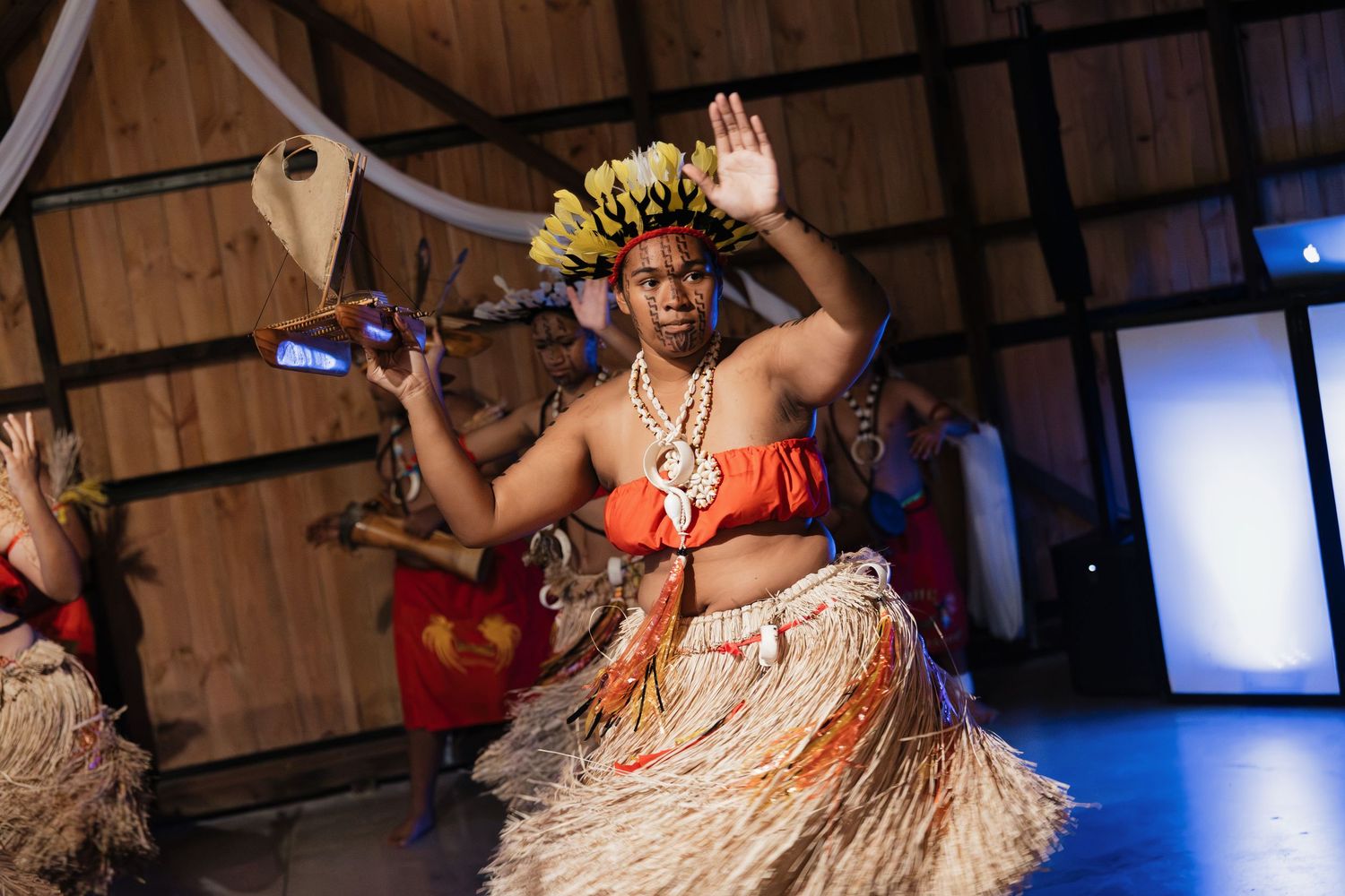 A Polynesian dancer in traditional grass skirt and floral headpiece performs gracefully at a cultural event indoors.