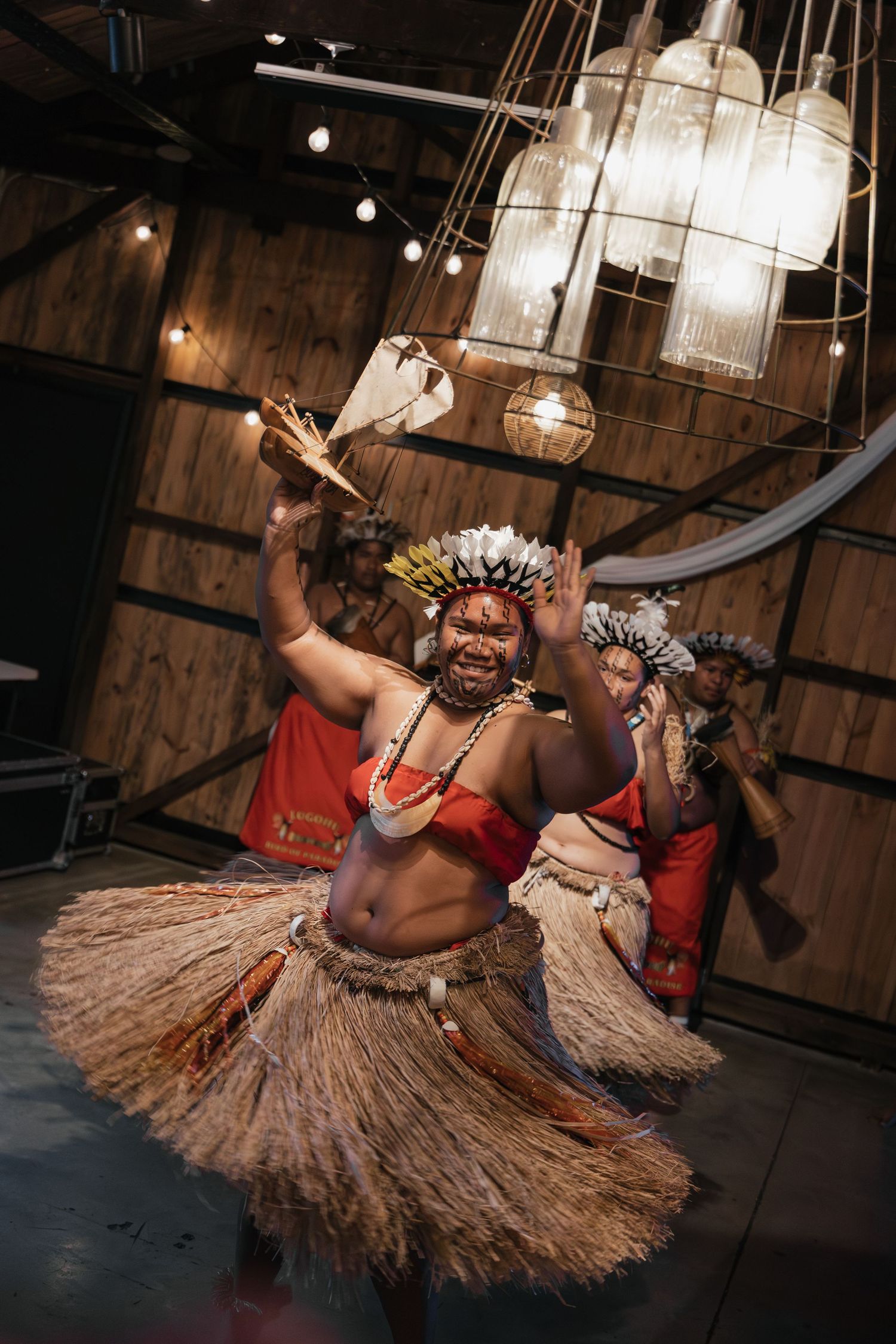 Traditional Pacific Islander dancers in grass skirts and headdresses performing energetically indoors under decorative lights.