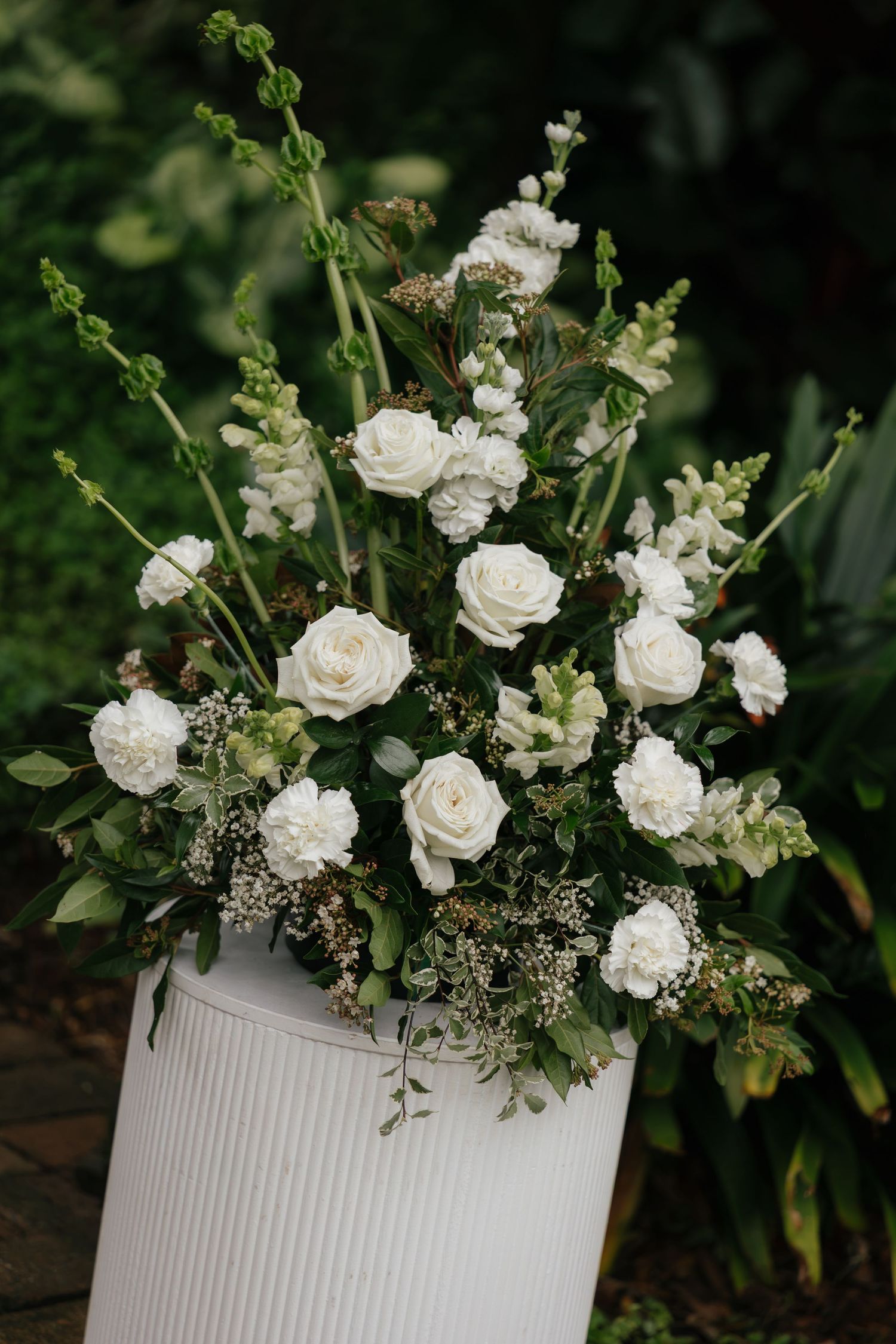 White roses and carnations arranged in a ribbed white vase with lush greenery outdoors.