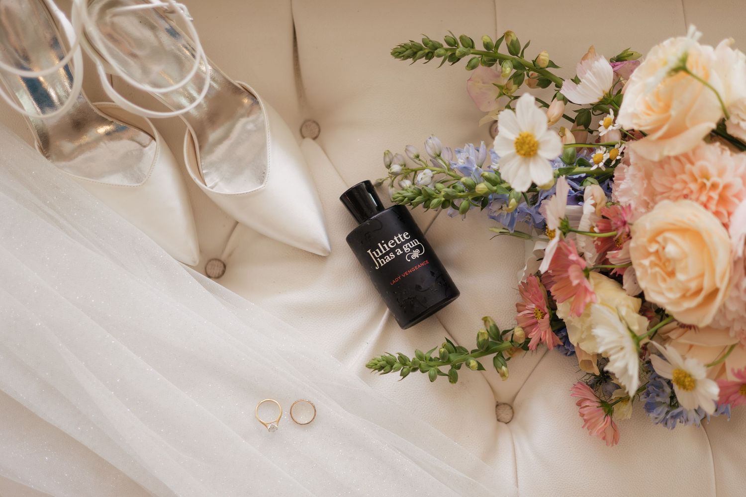 Wedding flat lay featuring bridal heels, a floral bouquet, gold rings, and Juliette Has a Gun perfume on white cushion.