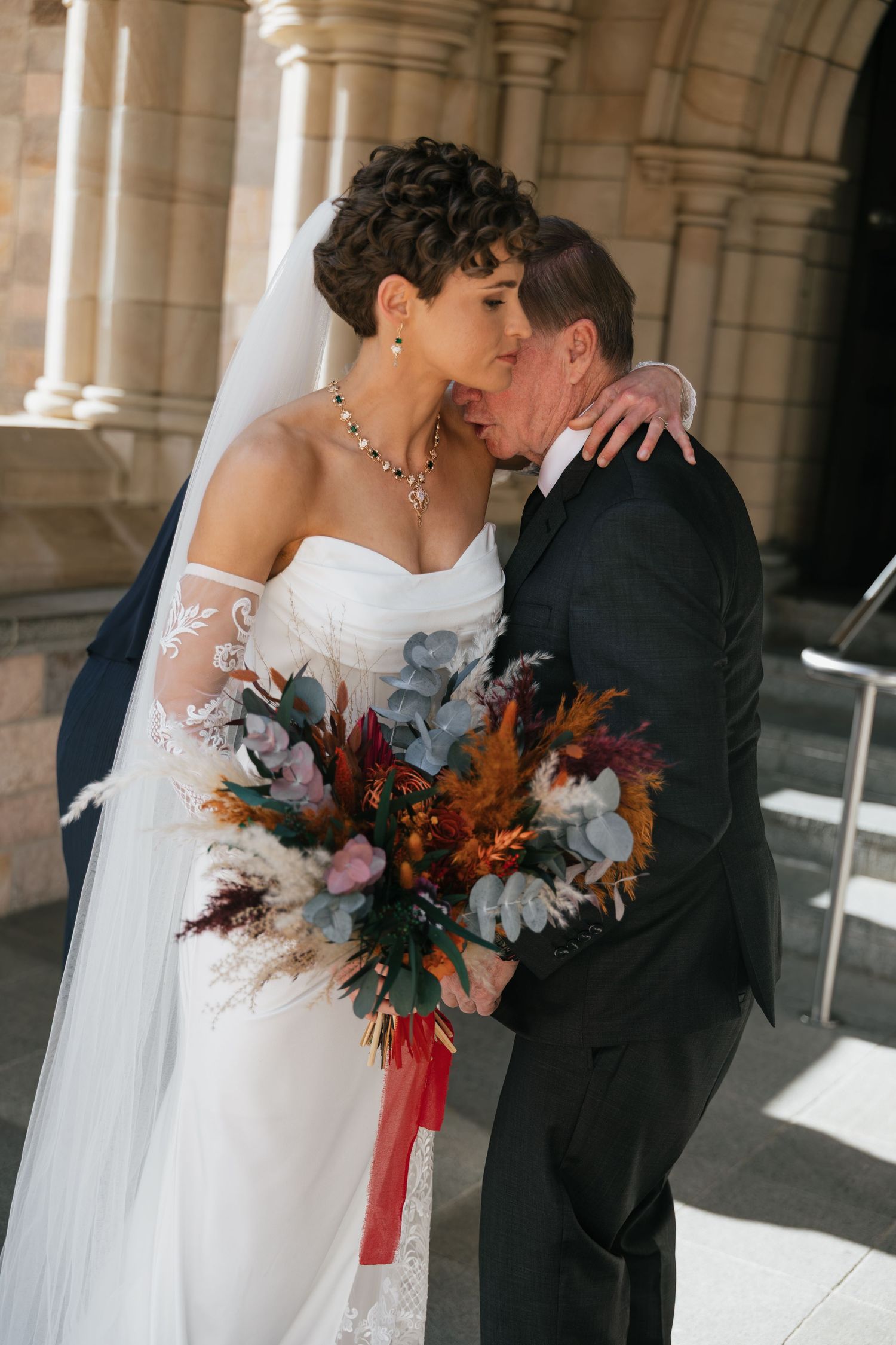 Bride and groom share a romantic kiss outside a church, bride holding a colorful fall bouquet in a white gown with veil.