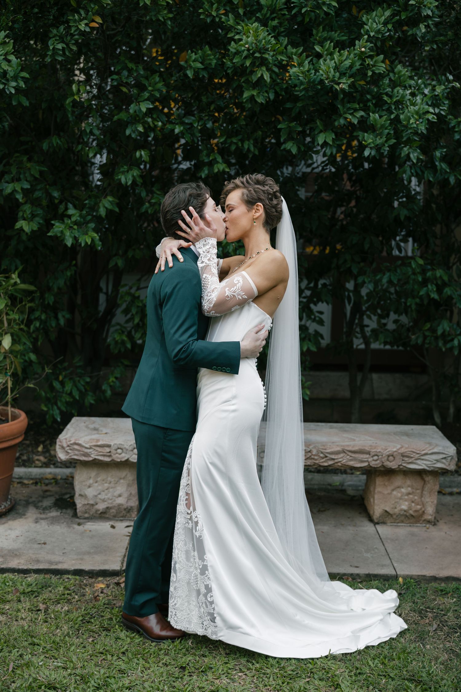 Bride and groom share a romantic kiss outdoors, bride wearing white gown with lace sleeves and veil, groom in green suit.