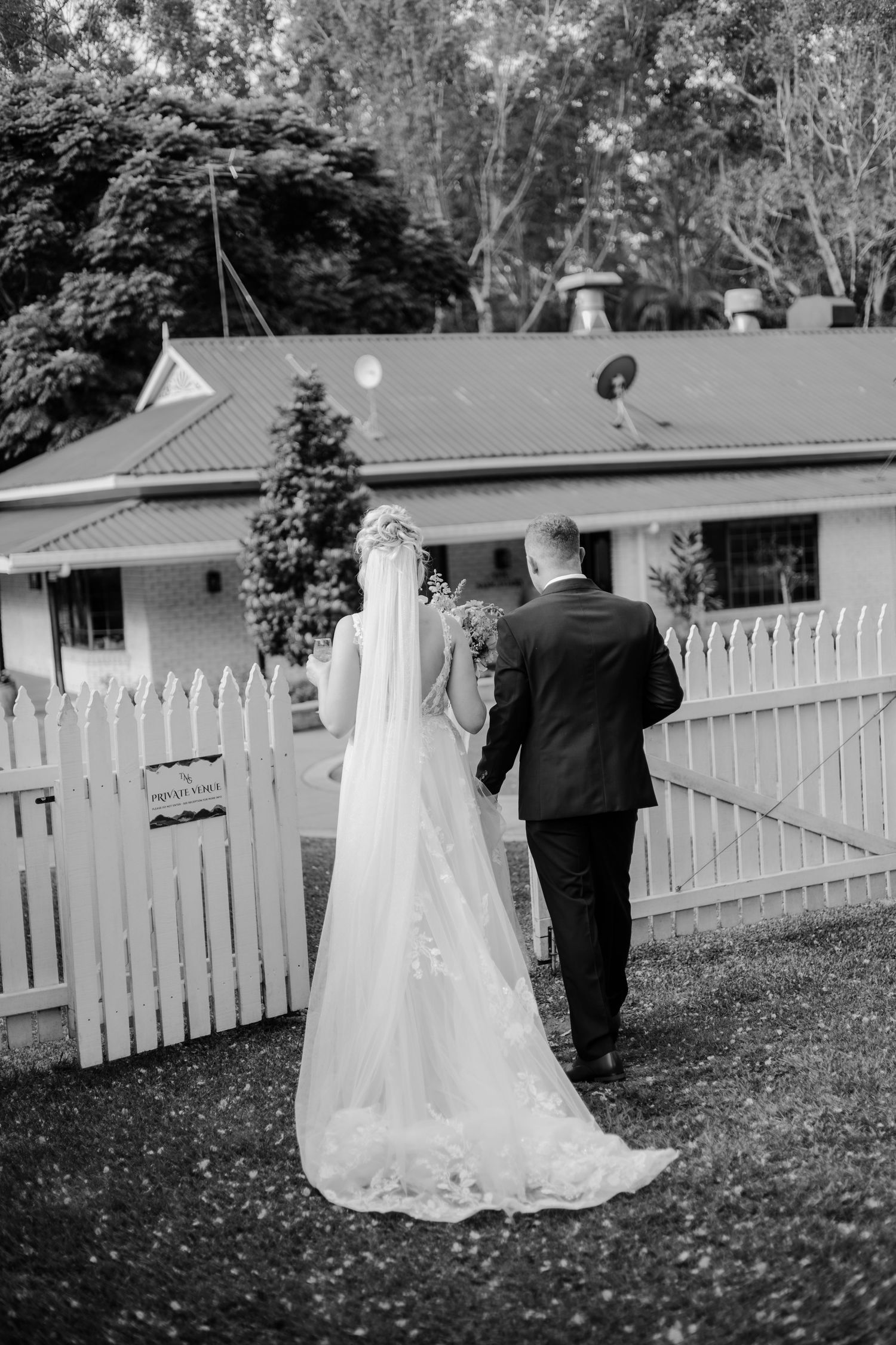 Black and white photo of a bride and groom walking hand in hand toward a white picket fence outside a rural home.