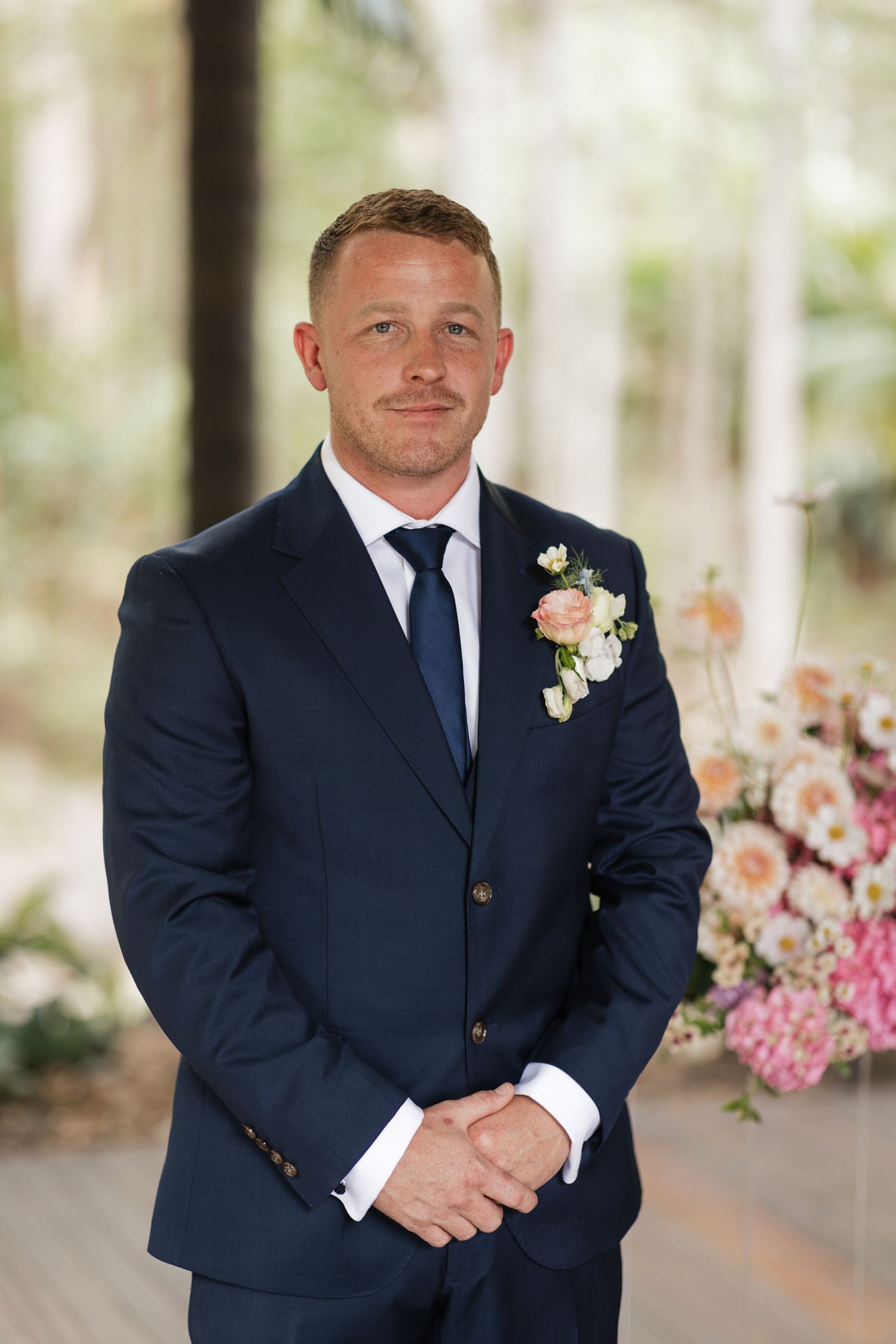 Groom in navy blue suit with floral boutonniere smiling at wedding ceremony with pink flowers in background.