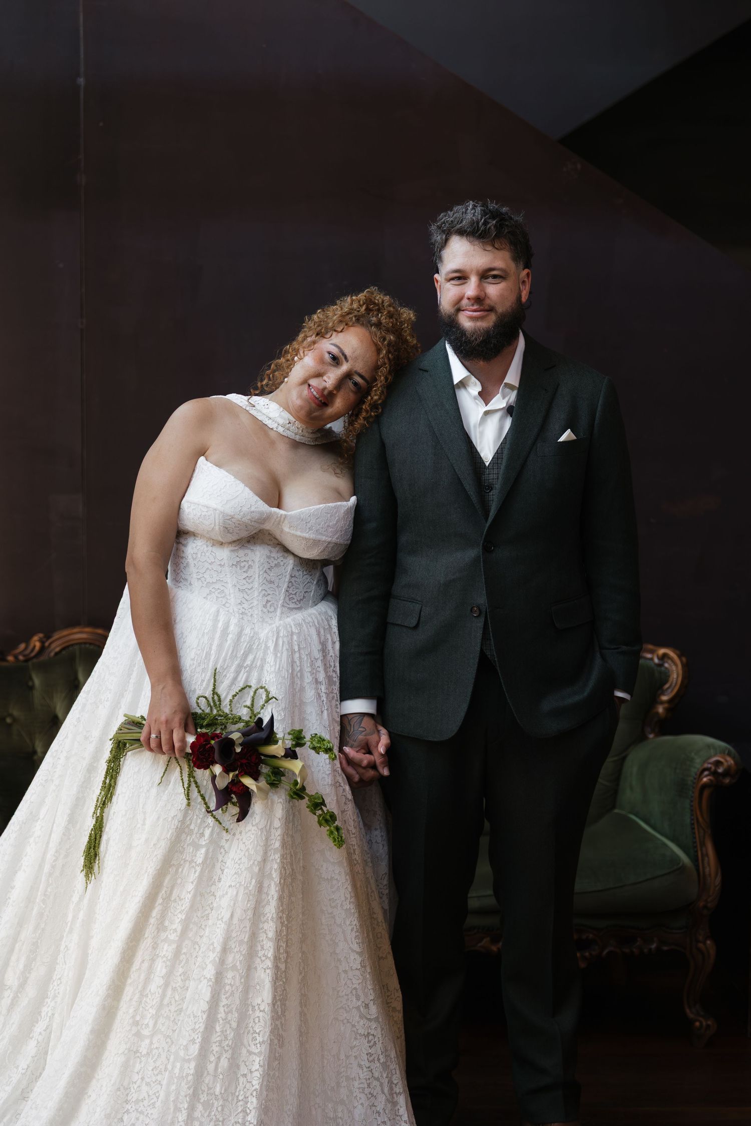 A bride in a white lace gown holds a bouquet while leaning on her groom in a dark green suit.
