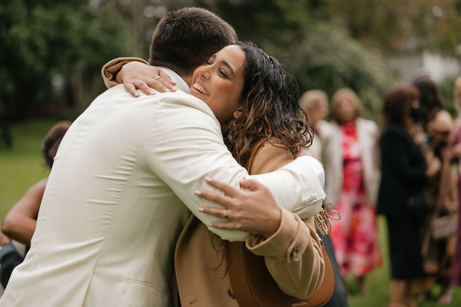 Two people share a warm embrace outdoors at a wedding celebration, with guests visible in the background.