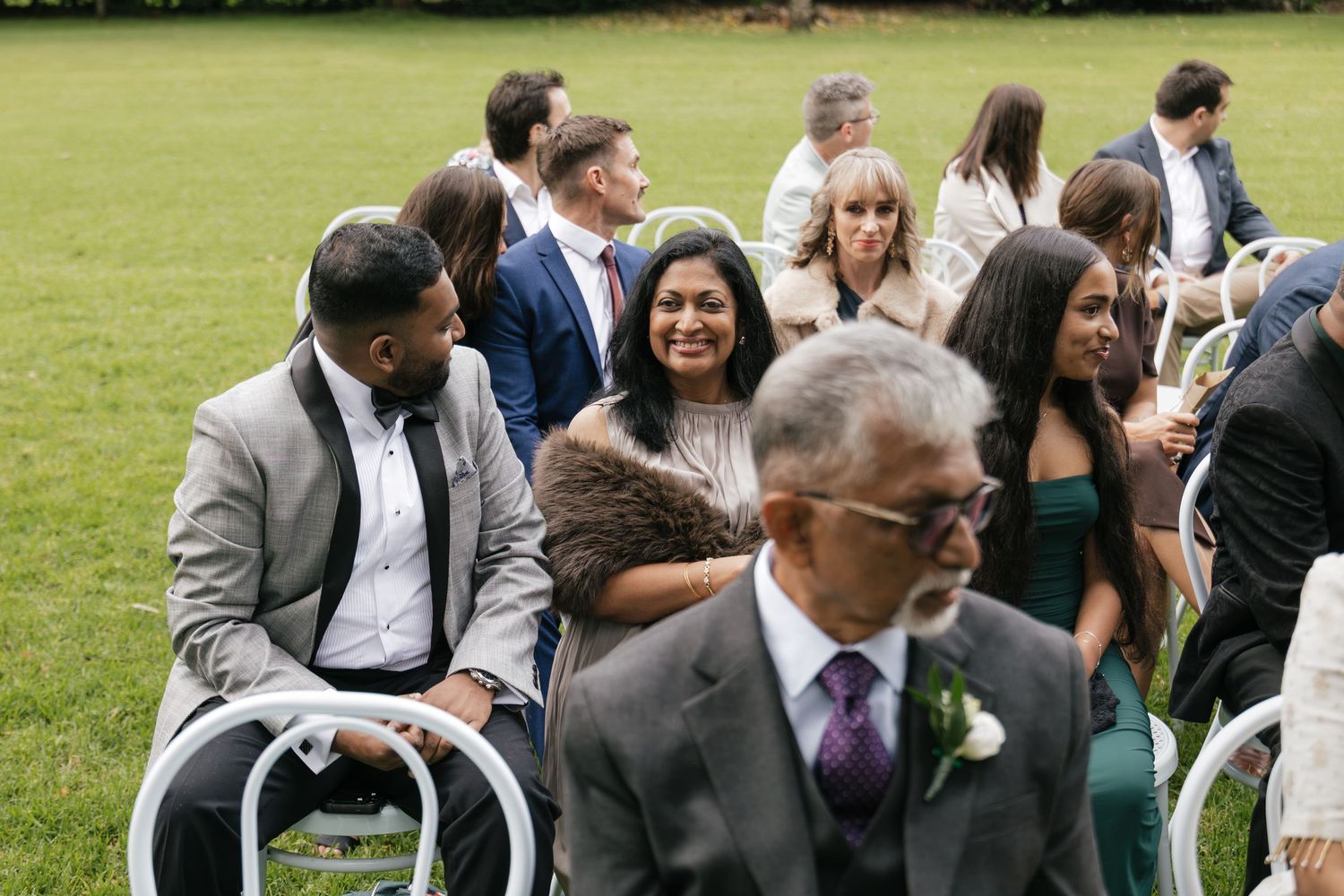 Guests smile and chat during an outdoor wedding ceremony seated on white chairs on a green lawn.