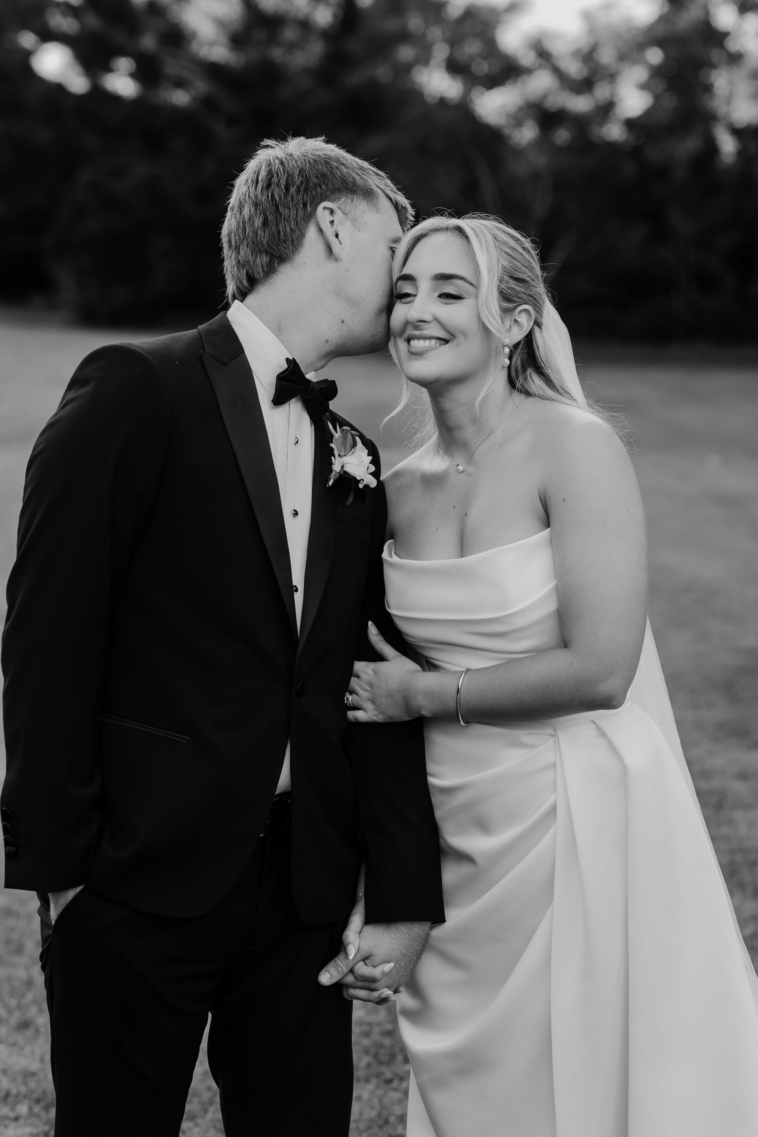 Black and white wedding photo of groom kissing bride on cheek, bride smiling in strapless gown, outdoors.