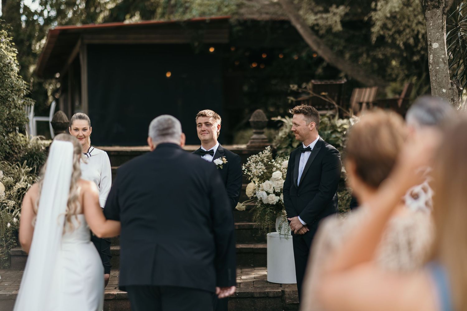 Outdoor wedding ceremony with bride, groom, and officiant at a rustic woodland venue with floral arrangements.