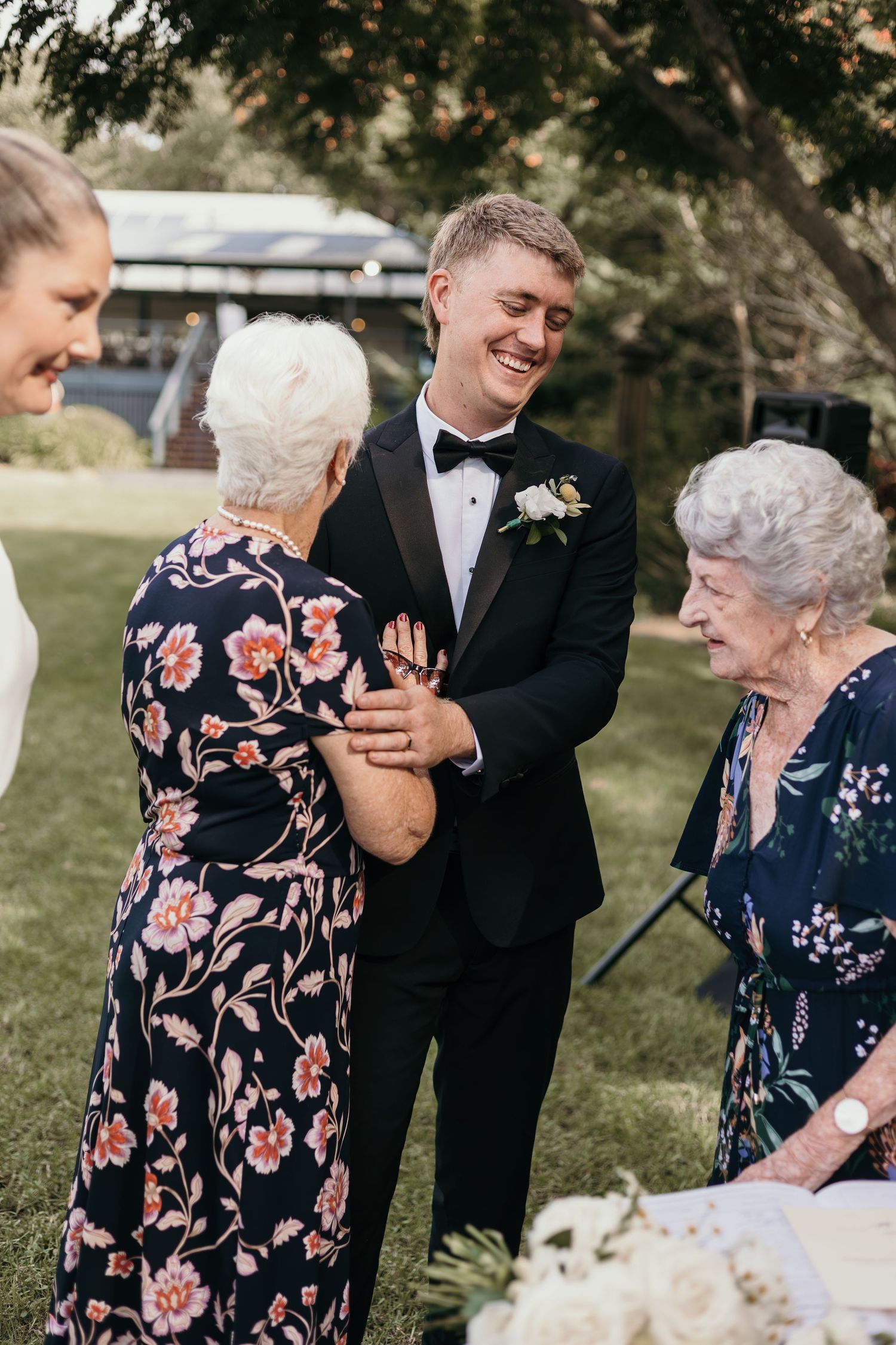 Groom in tuxedo laughing with elderly women in floral dresses at an outdoor wedding reception.