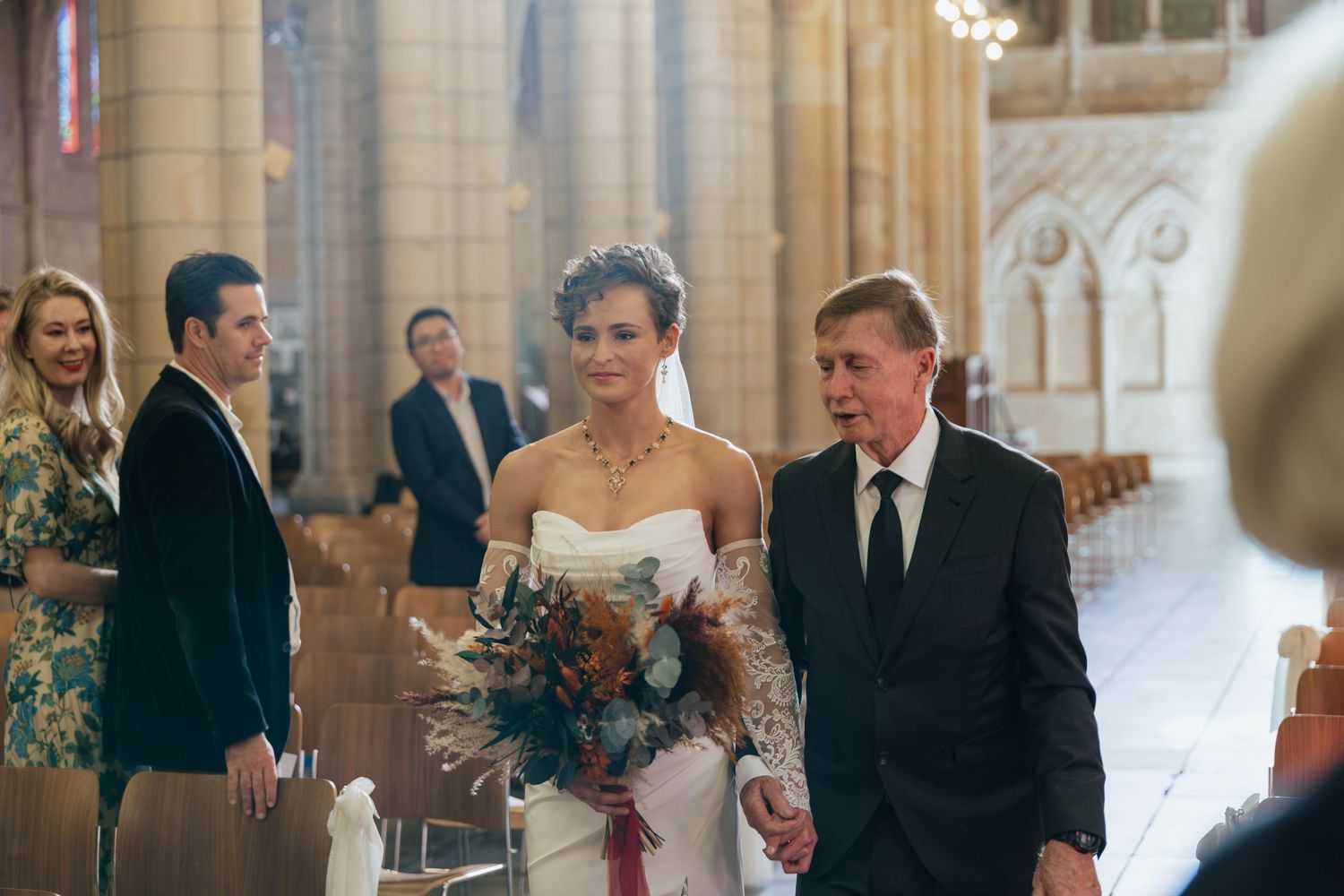 Bride walks down church aisle with older man, holding bouquet, guests watching in elegant stone cathedral.