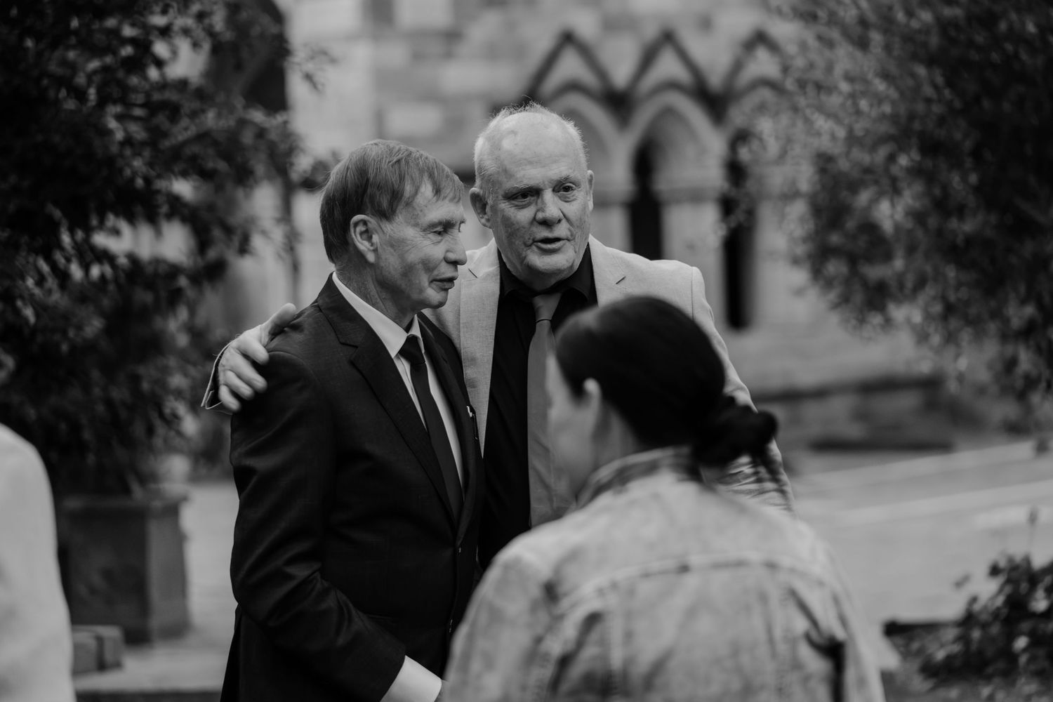 Two men in formal attire embrace outdoors near a historic stone church in a black and white photo.