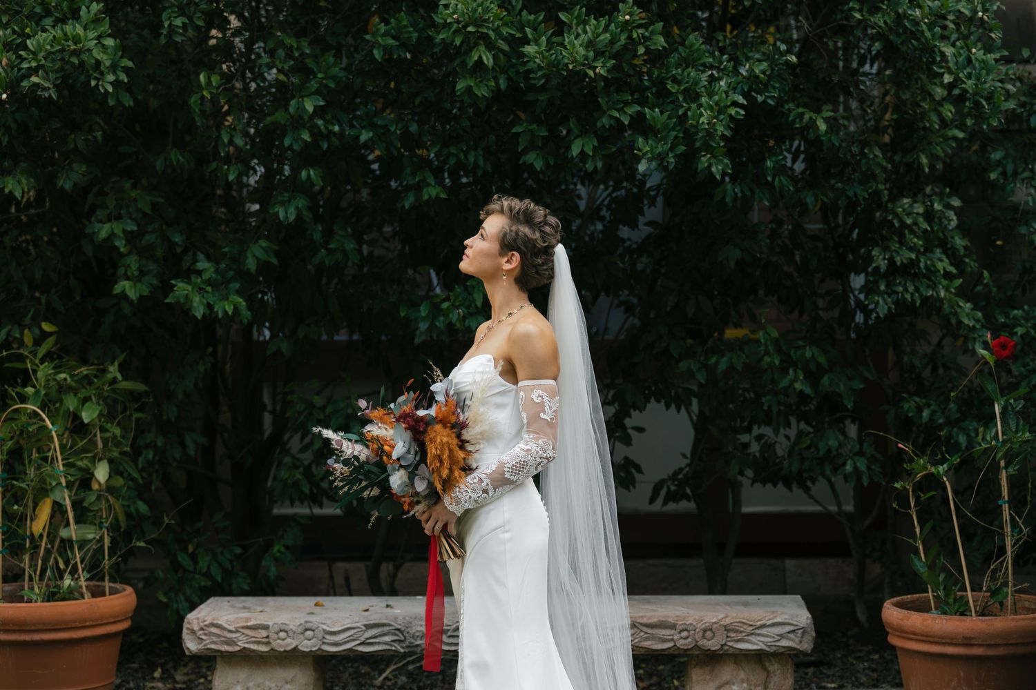 Bride in white off-shoulder gown with veil holds bouquet in lush garden setting.