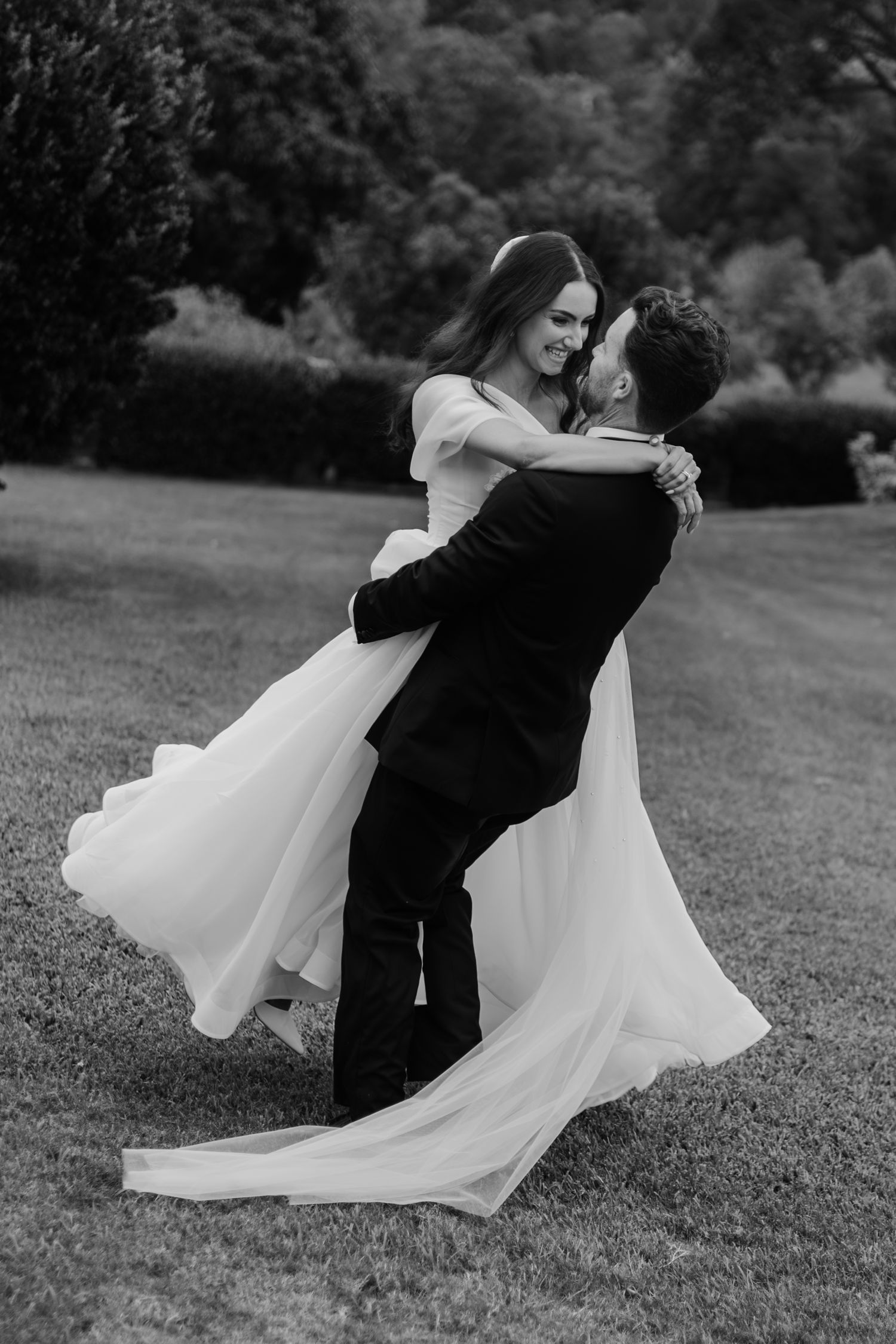 Black and white photo of groom lifting bride in flowing wedding dress on lush green lawn.