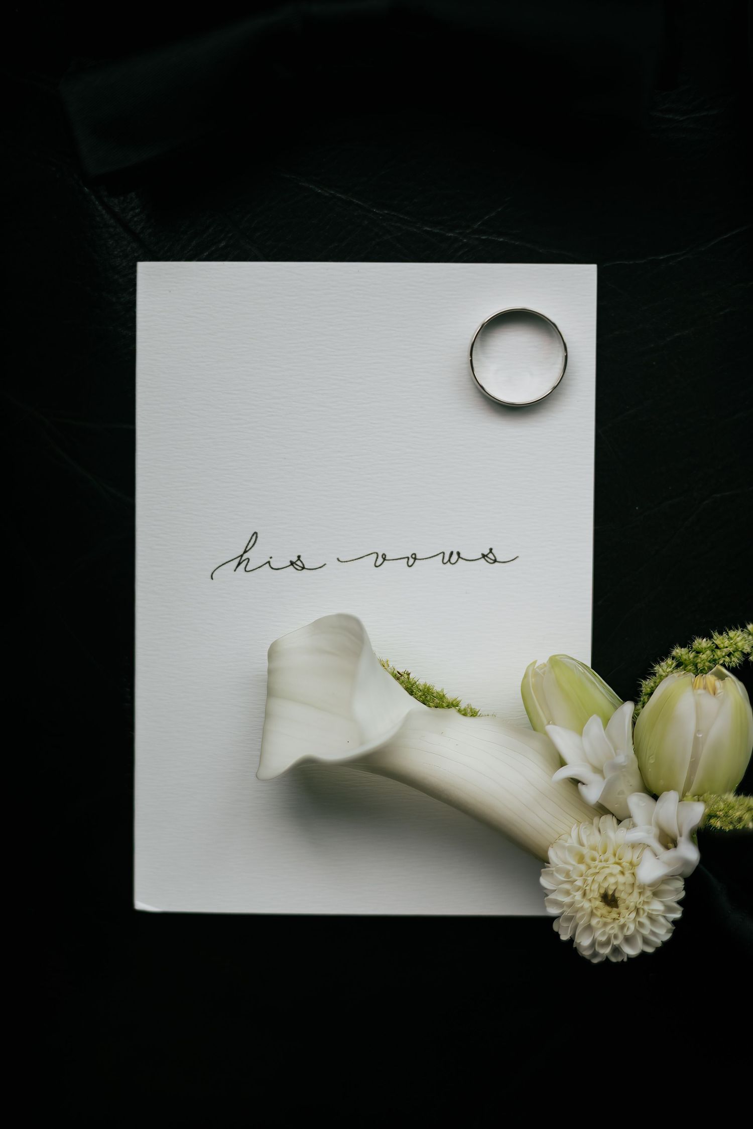 A wedding vow card reading his vows with a silver ring and white calla lily flowers on a dark green background.