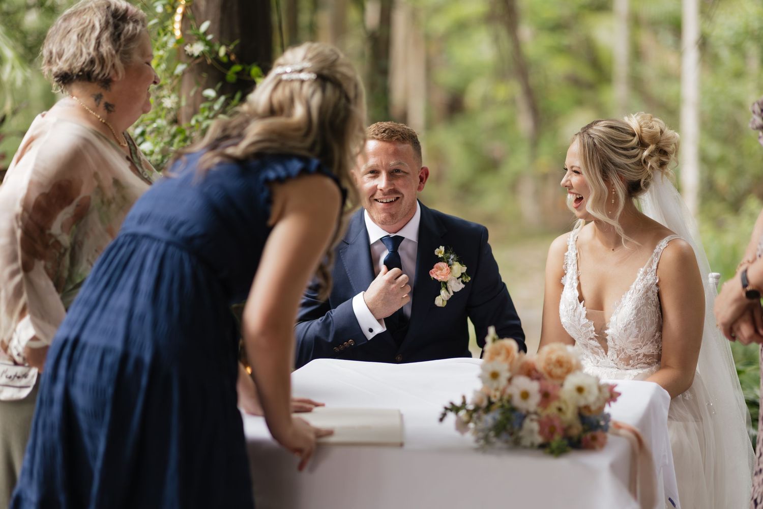 Bride and groom smile at wedding table with guests in navy dress, surrounded by floral arrangements in outdoor forest setting.