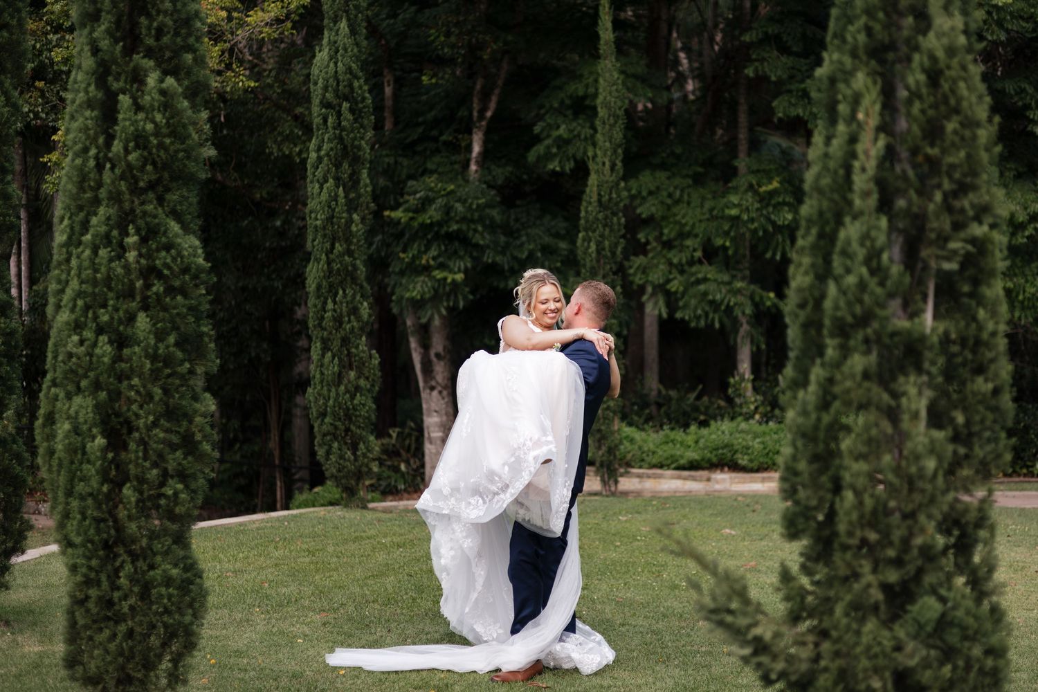 Groom lifting bride in white gown among tall cypress trees on lush green lawn at outdoor wedding.