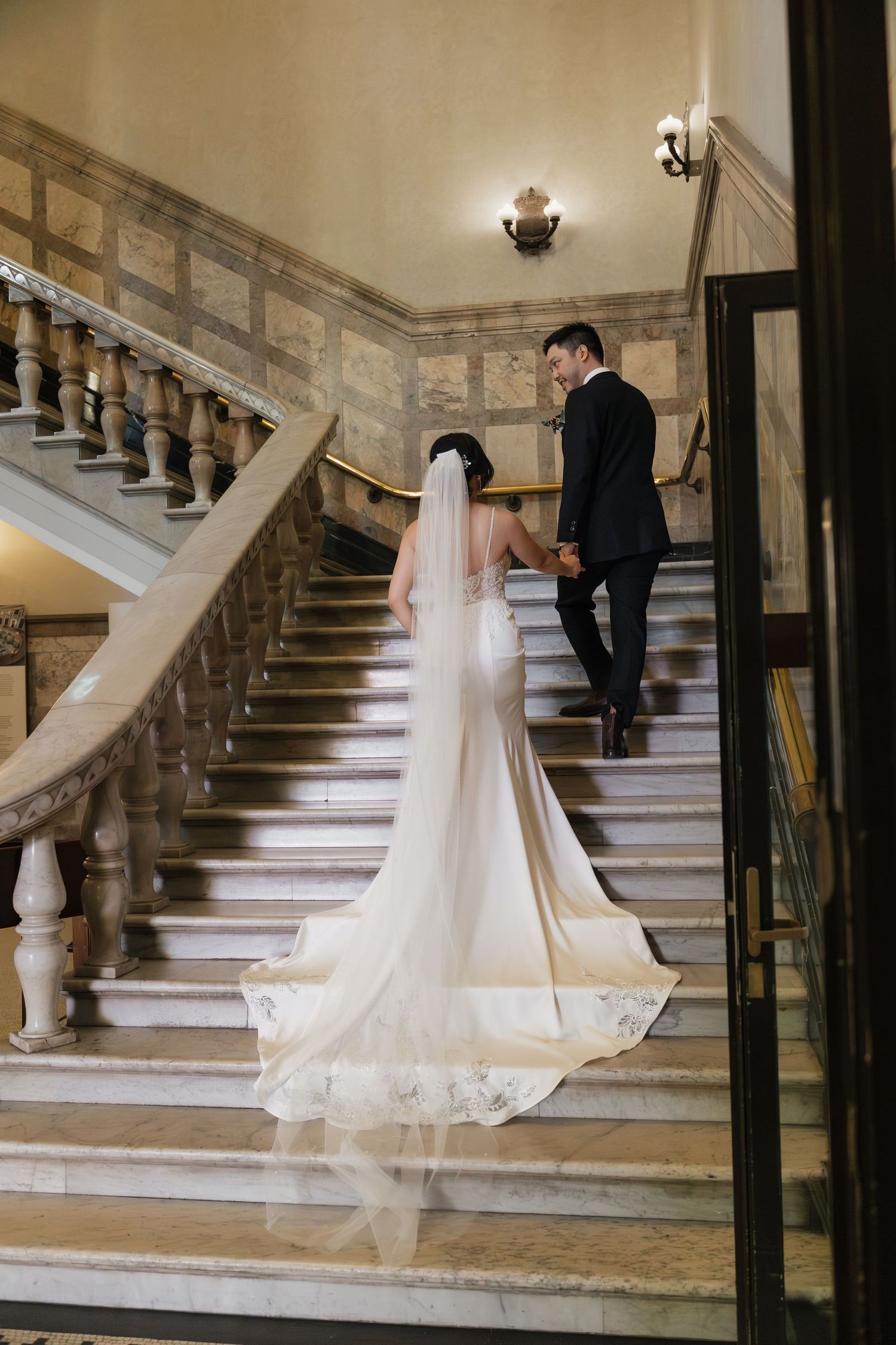 Bride and groom walking up elegant marble staircase, bride wearing long white gown with flowing veil and train.