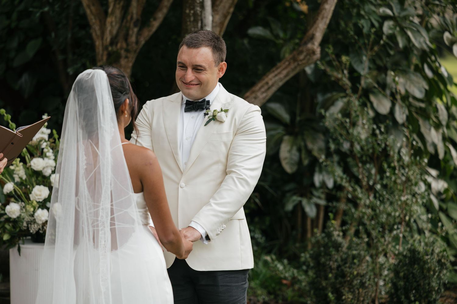 Groom in white tuxedo jacket smiling at bride in wedding veil during outdoor ceremony with greenery backdrop.