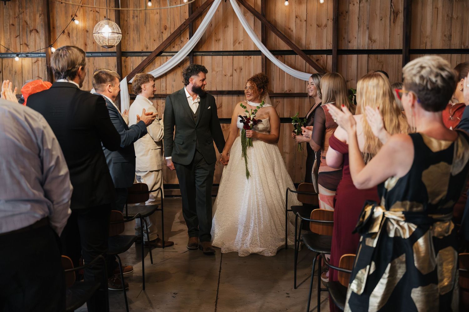 Bride and groom walk down the aisle in a rustic barn wedding venue as guests applaud and celebrate.