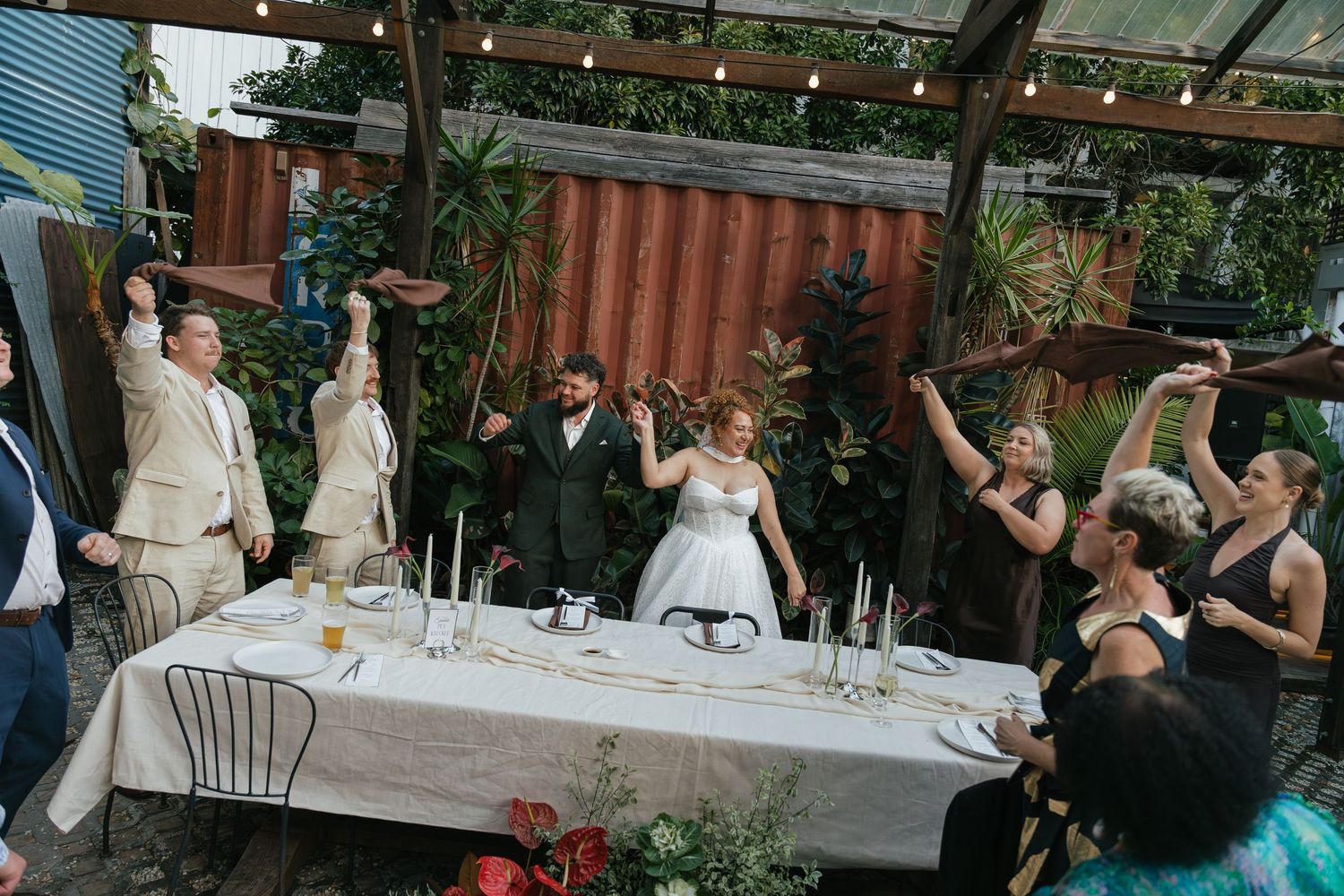 Wedding party raises glasses in toast at outdoor reception table surrounded by lush greenery and string lights.