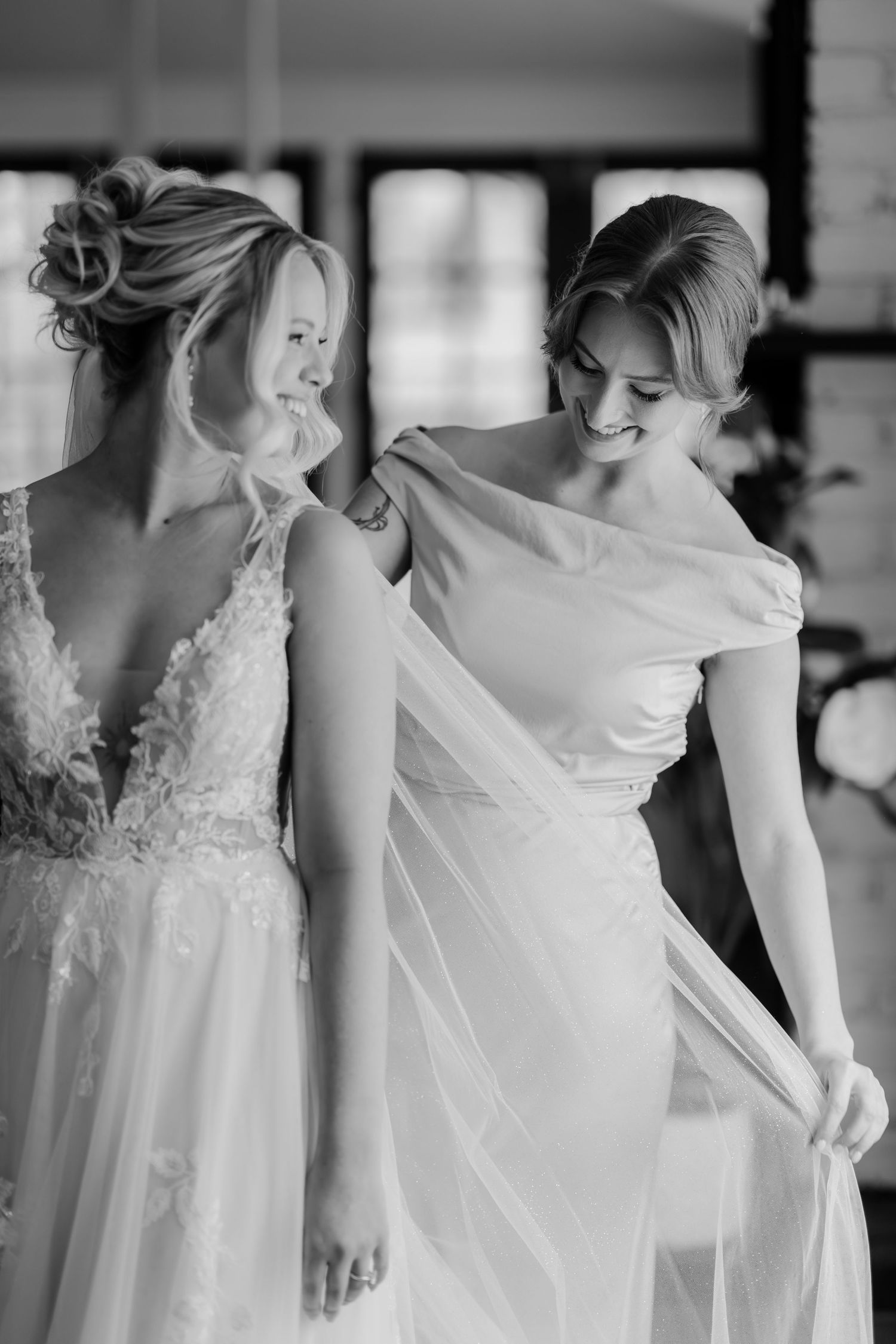 Black and white photo of a bride in lace gown as smiling bridesmaid adjusts her flowing veil indoors.