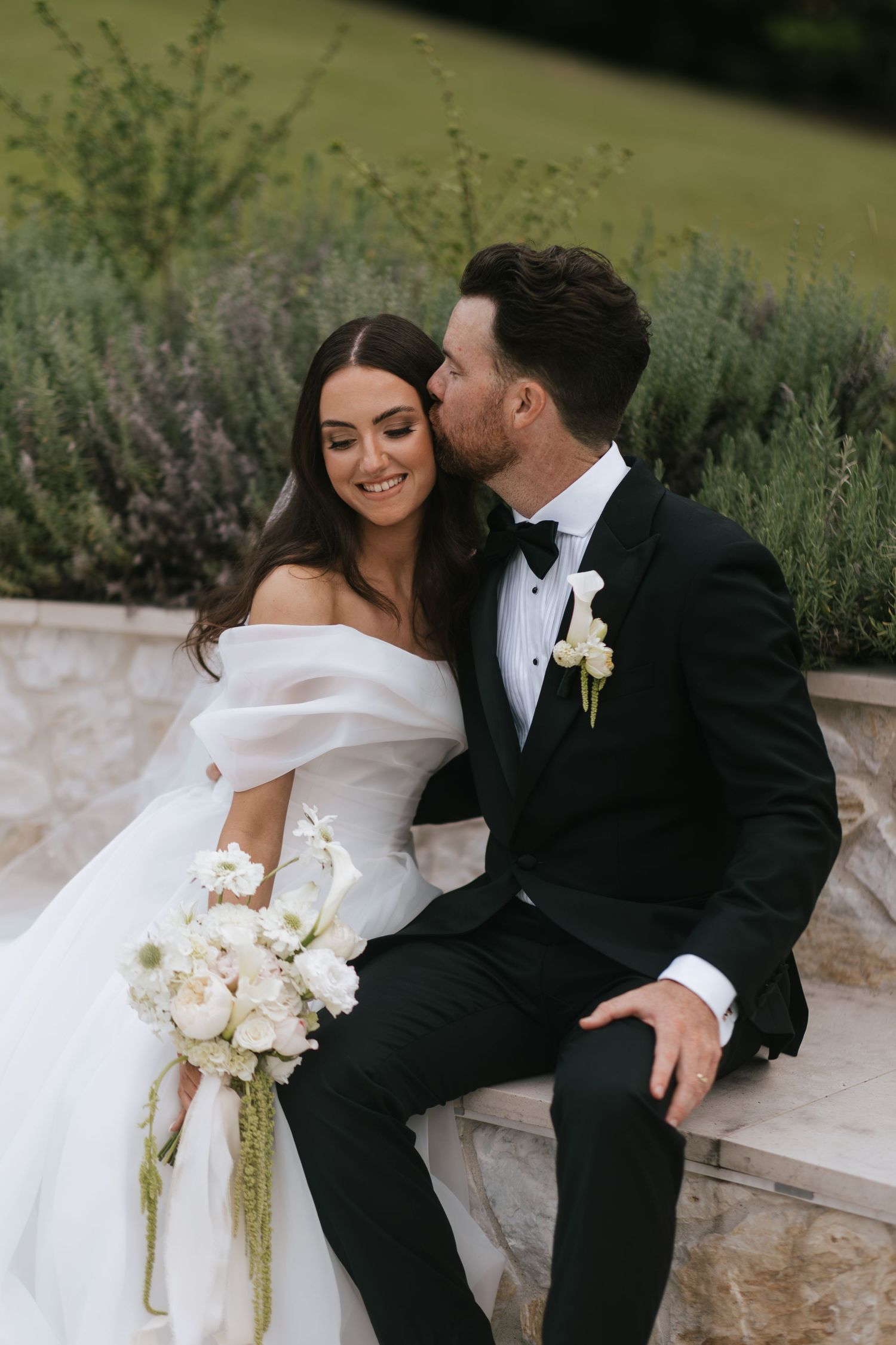 Bride and groom sitting outdoors, groom kissing bride's cheek, bride holding white floral bouquet in elegant off-shoulder gown.
