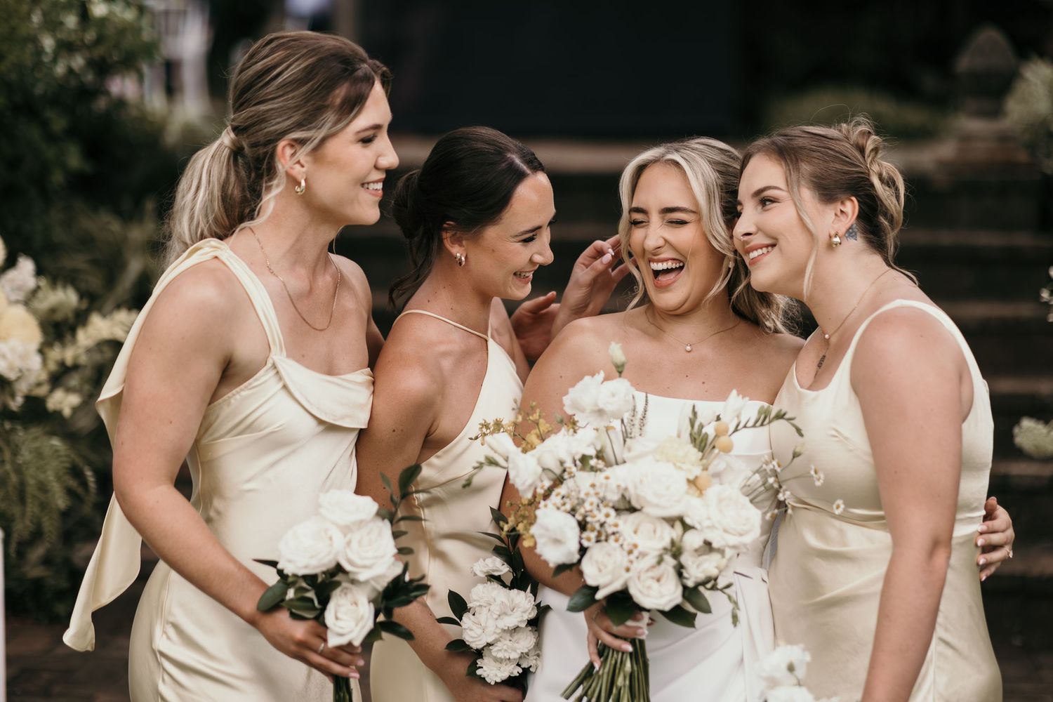 Four bridesmaids in ivory satin dresses laughing together, holding white floral bouquets at an elegant wedding.